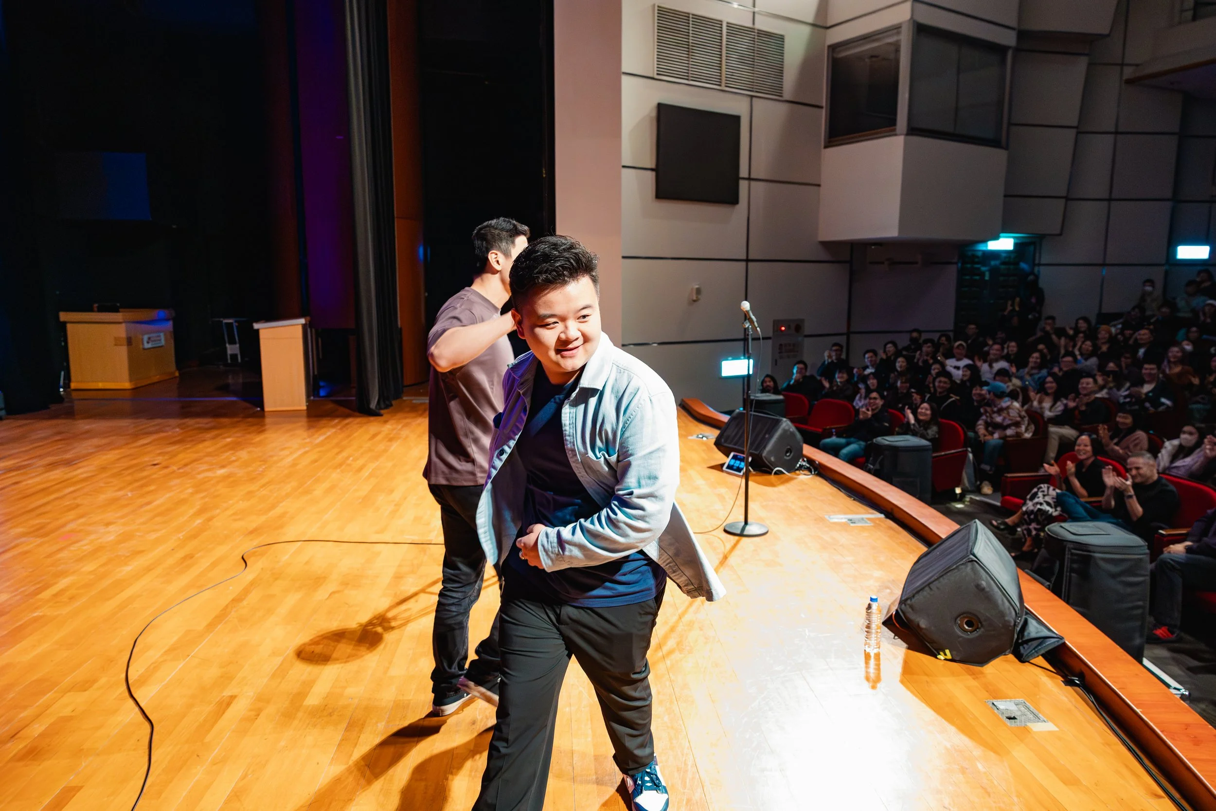 Two young men on stage, one sitting and smiling while the other stands behind him, in front of a large audience in an auditorium.