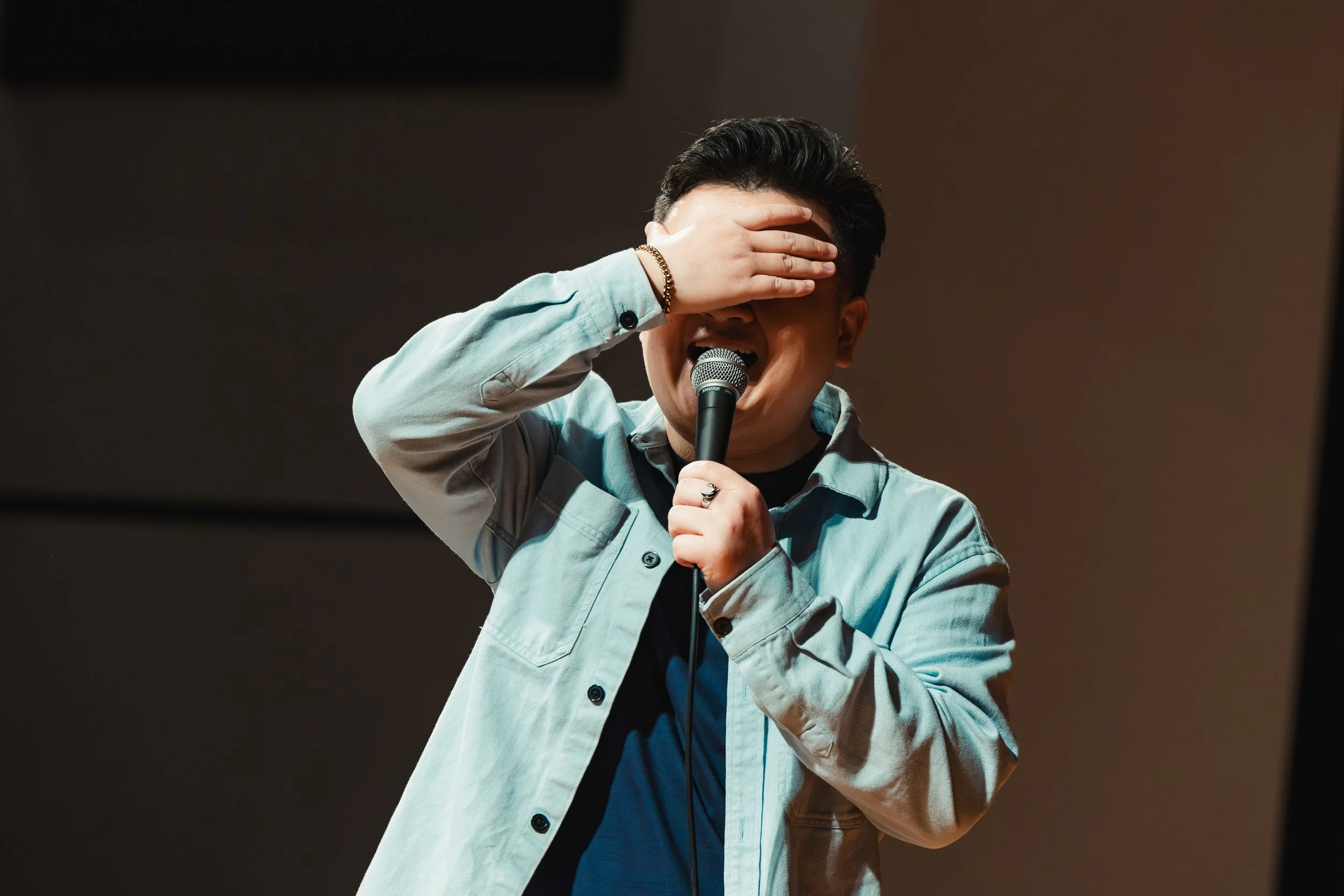 A young man with dark hair wearing a light-colored jacket and holding a microphone, with his hand covering his face in front of a dark background.