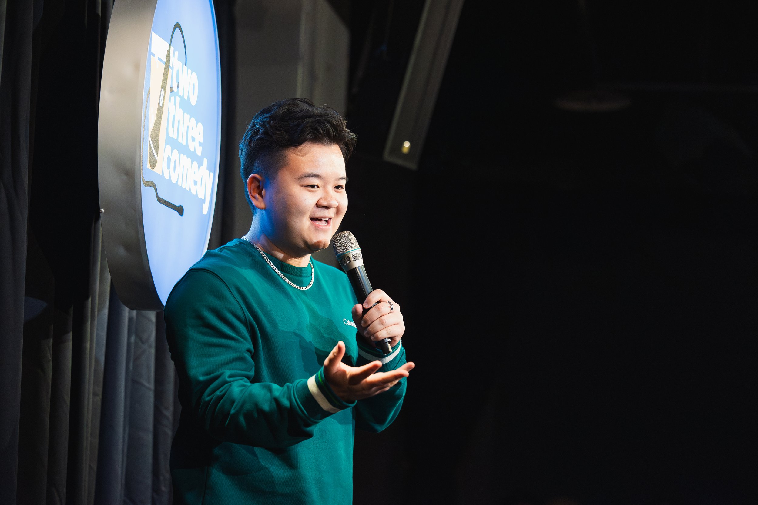 A young man smiling and speaking into a microphone on stage during a comedy show. He is wearing a teal long-sleeve shirt and a silver chain, with a comedy sign in the background.