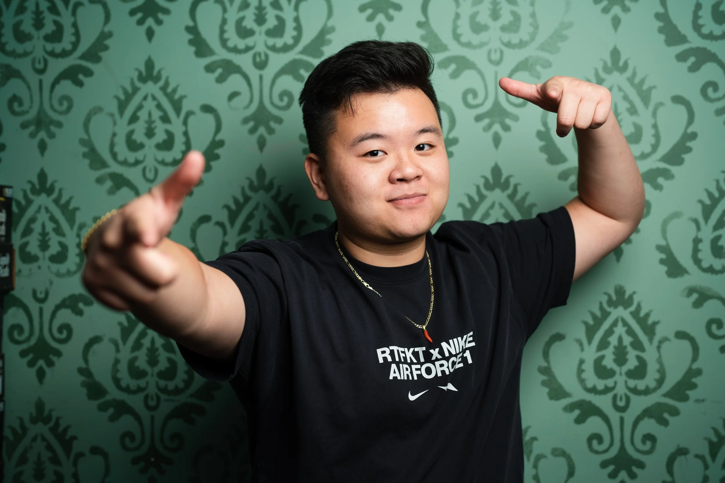 Young man with black hair wearing a black Nike t-shirt and a necklace, pointing towards the camera with both hands, standing in front of a green patterned wallpaper.