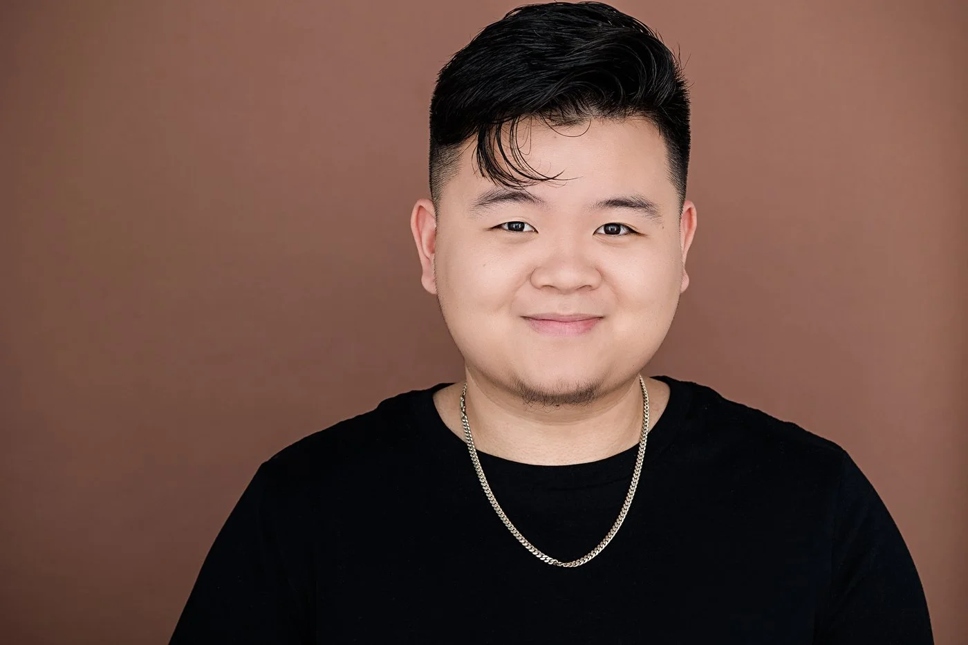 A young man with short black hair, light skin, and a goatee, smiling, wearing a black shirt and a silver chain necklace, against a brown background.
