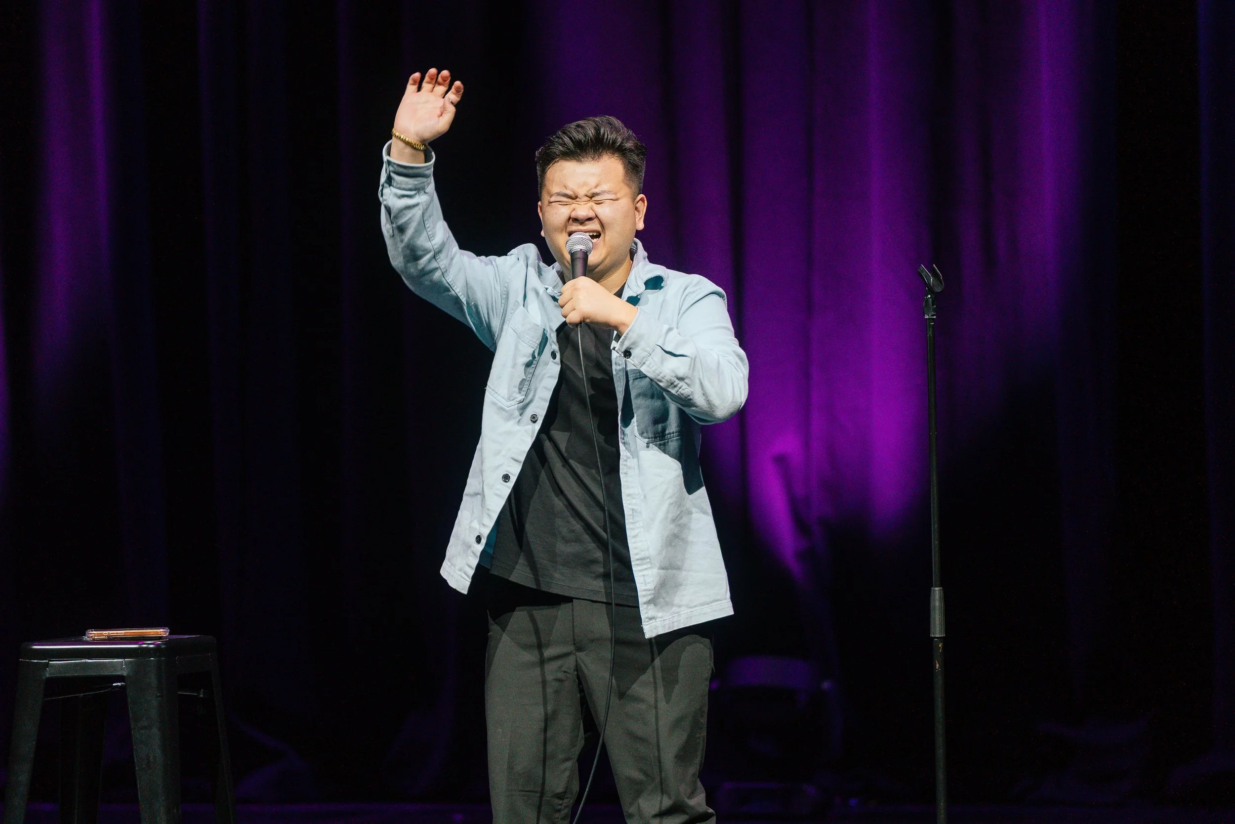 A man performing stand-up comedy on stage. He has short dark hair, is wearing a light-colored jacket over a dark shirt, and is holding a microphone with his eyes closed and an expressive face. The stage has purple curtains in the background, and a bl