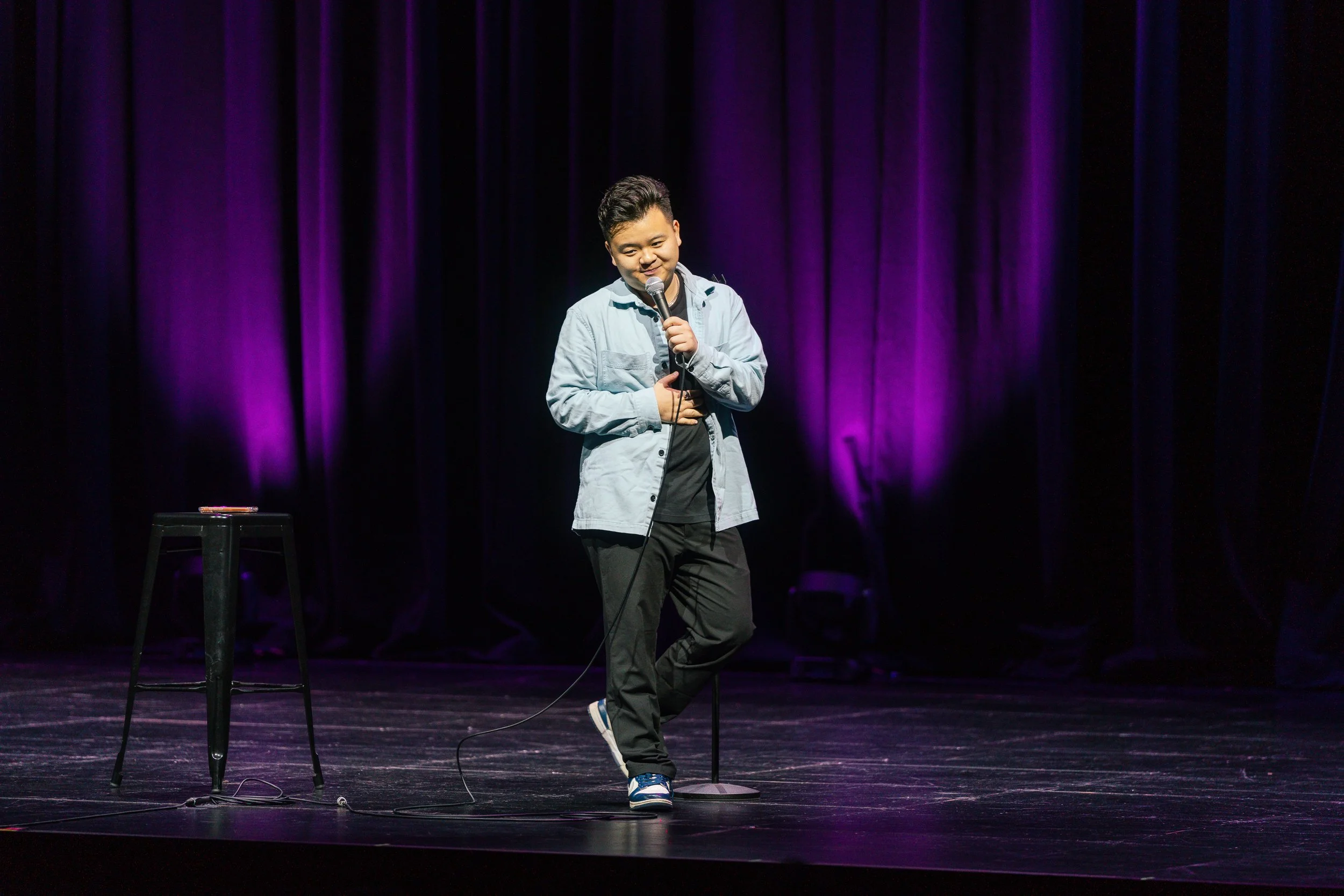 A young man standing on a stage holding a microphone, with a purple curtain backdrop, a stool with papers nearby, dressed in casual clothes, smiling.