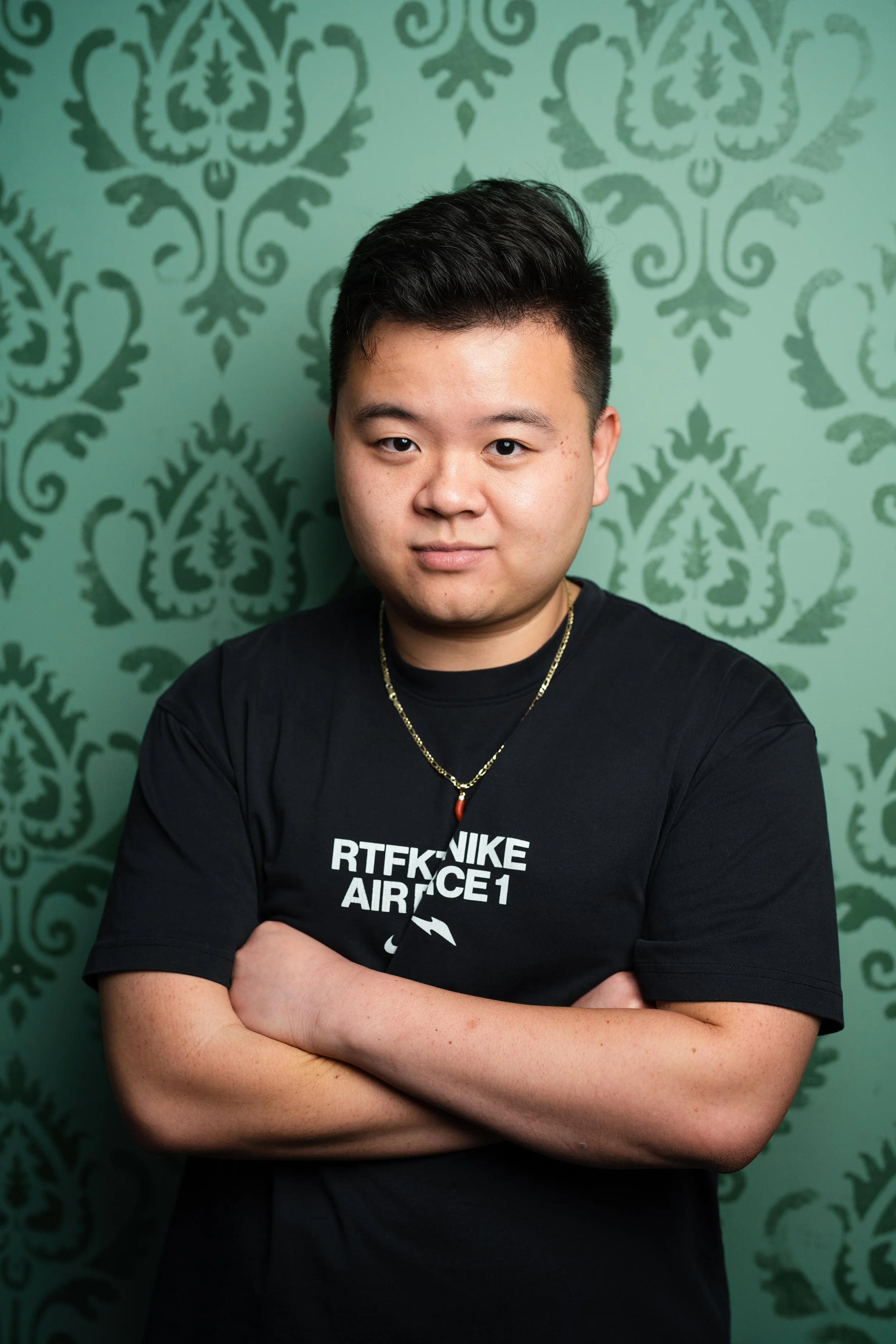 A young man with short black hair and light skin, standing with his arms crossed in front of a green patterned background, wearing a black T-shirt with white text and gold chains around his neck.