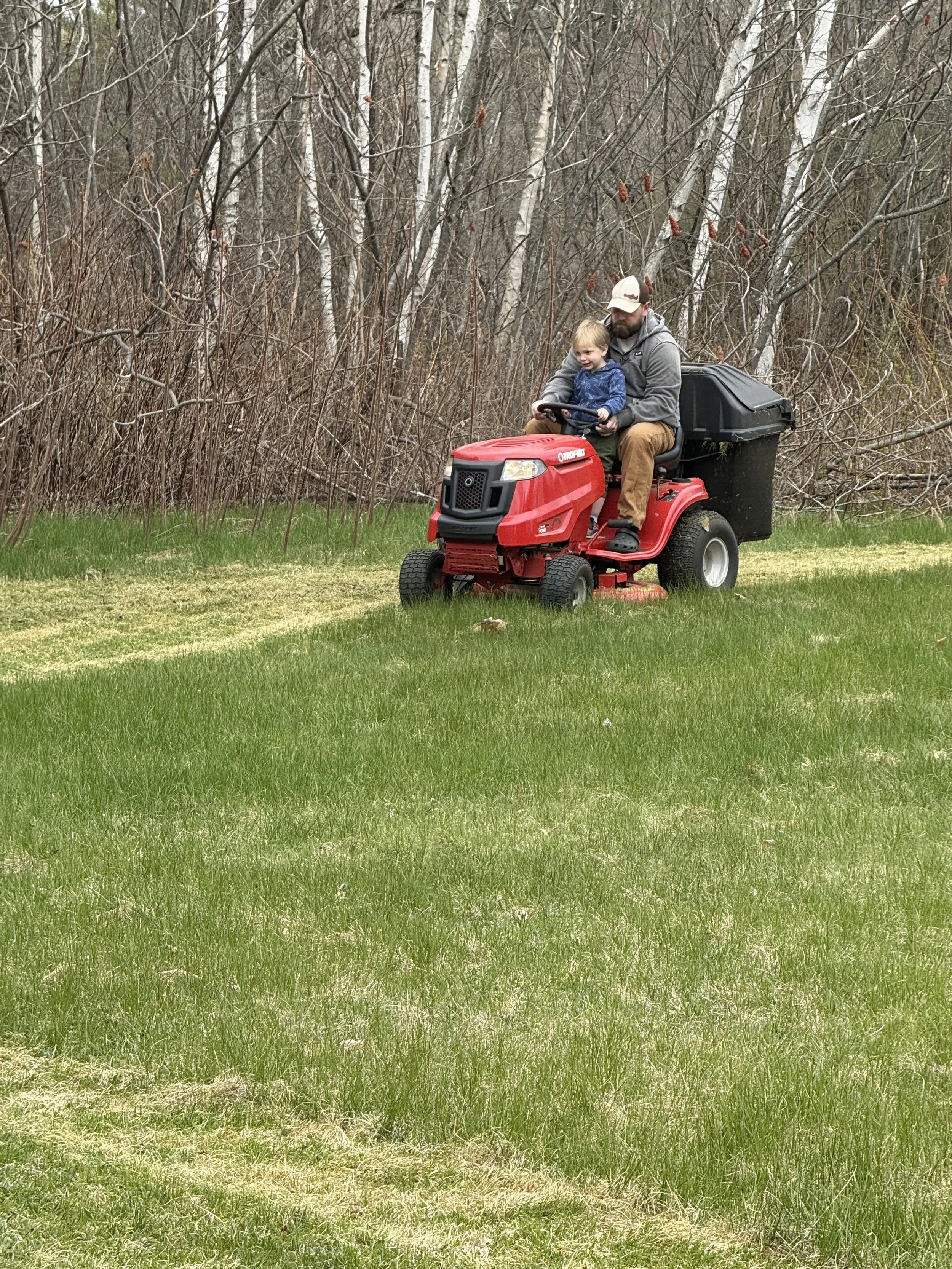 A man and a young child riding a red riding lawn mower across a grassy lawn with a background of bare trees and shrubs.