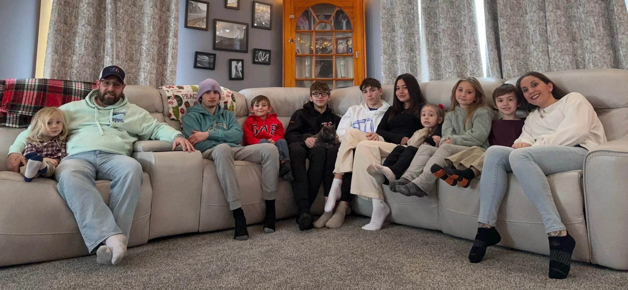 Family sitting on a couch in a living room, smiling at the camera. The group includes children and adults, with a small black cat on one person's lap. The cozy setting features curtains, framed pictures, and a decorative wooden window in the background.