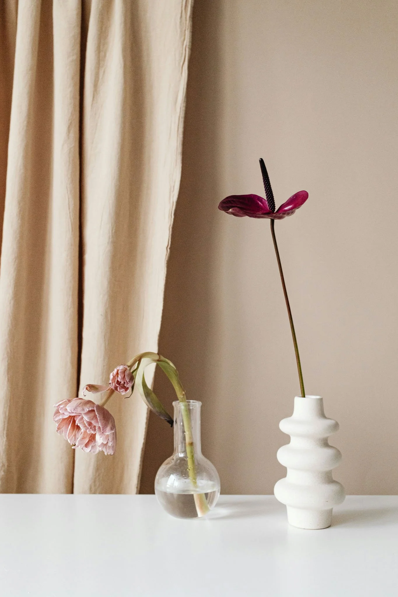 A beige curtain hanging beside a table with a white surface, holding two vases with dried flowers. One vase is clear glass with a single long-stemmed flower, the other is a white ceramic vase with a layered, geometric design.