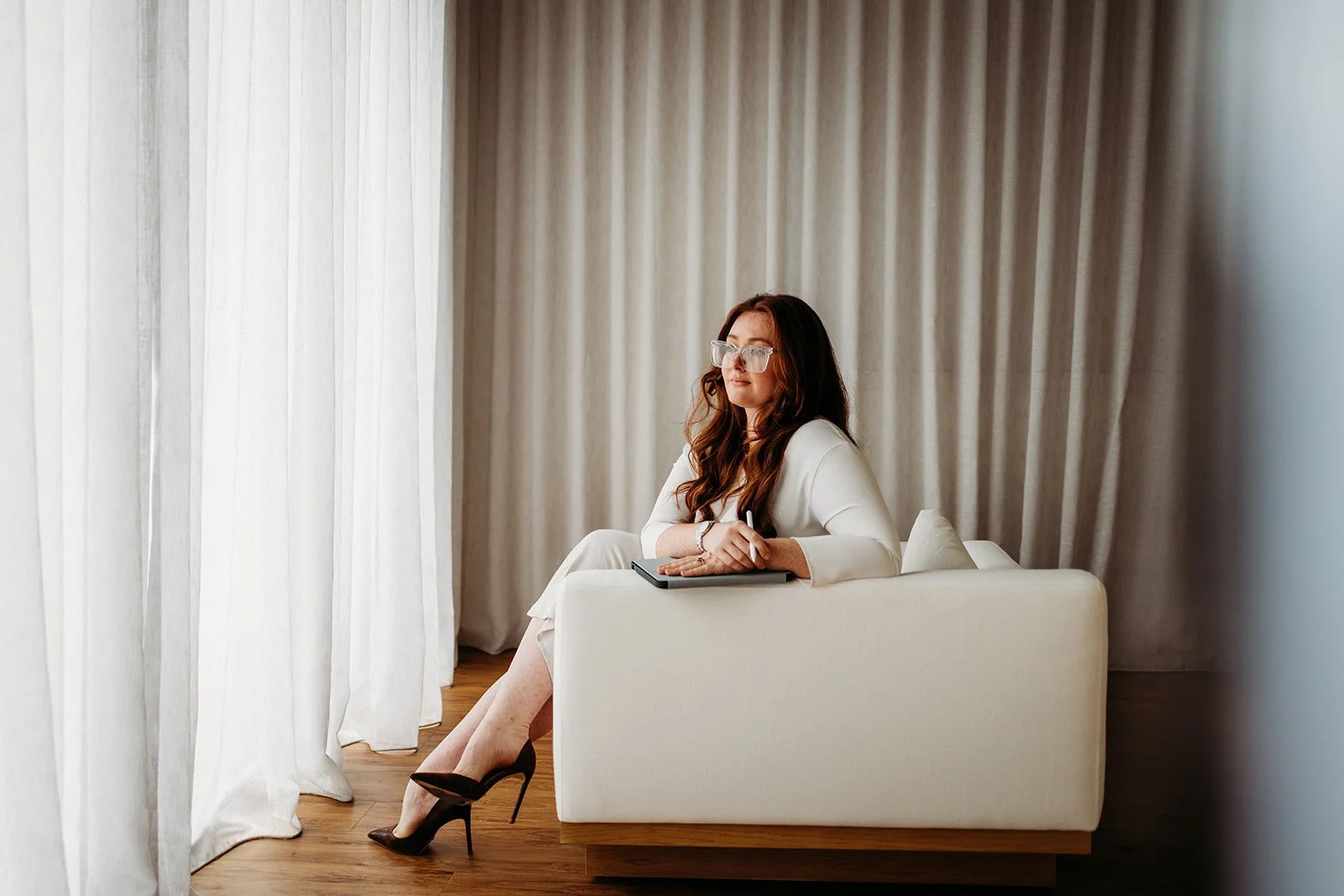 A woman with long brown hair and glasses, dressed in a white blazer and skirt, sitting on a white couch with her legs crossed, holding a stylus and notepad, looking out a window with white curtains.