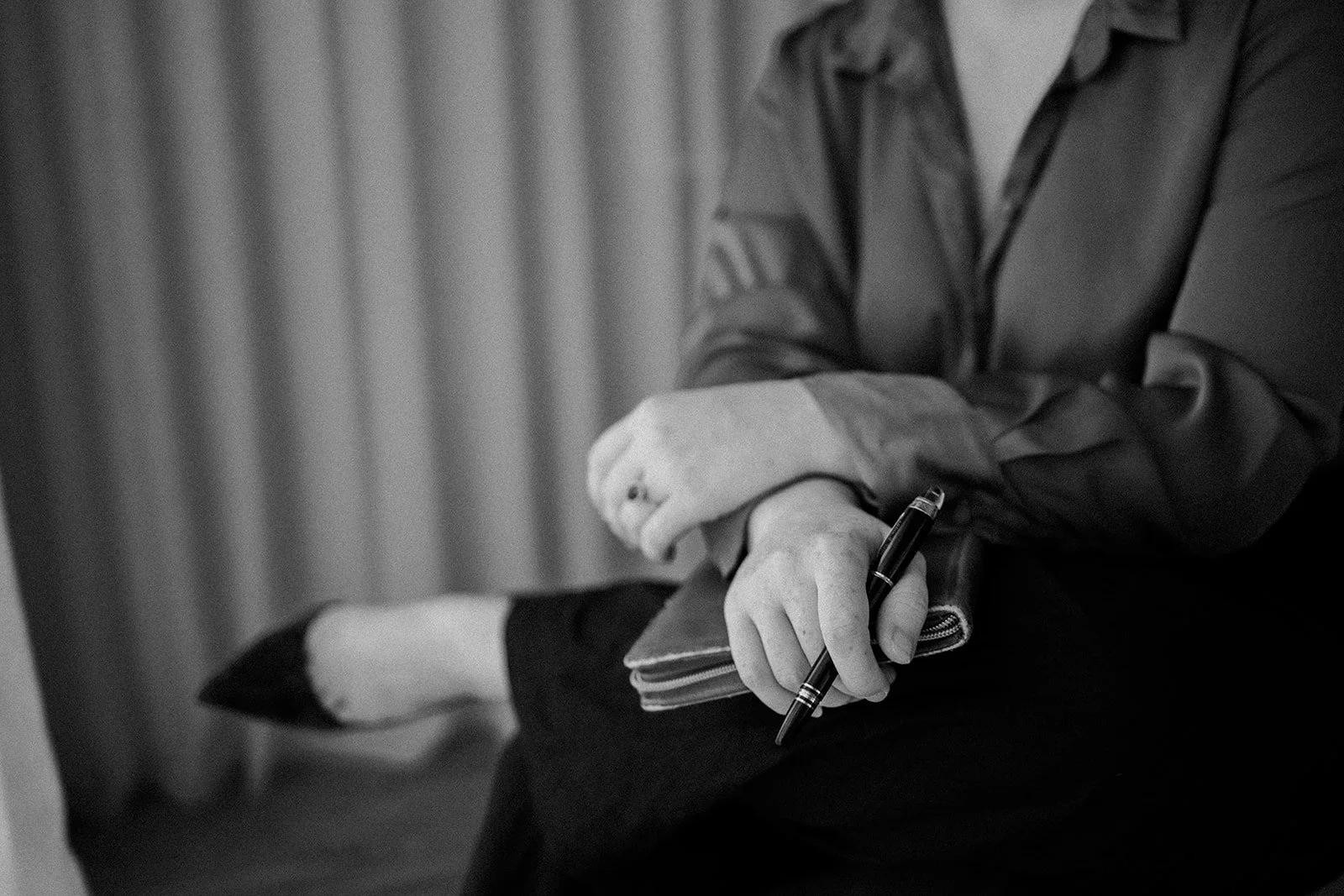 A person sitting with arms crossed, holding a pen and a notebook, wearing a button-up shirt and sitting on a chair with a curtain in the background.