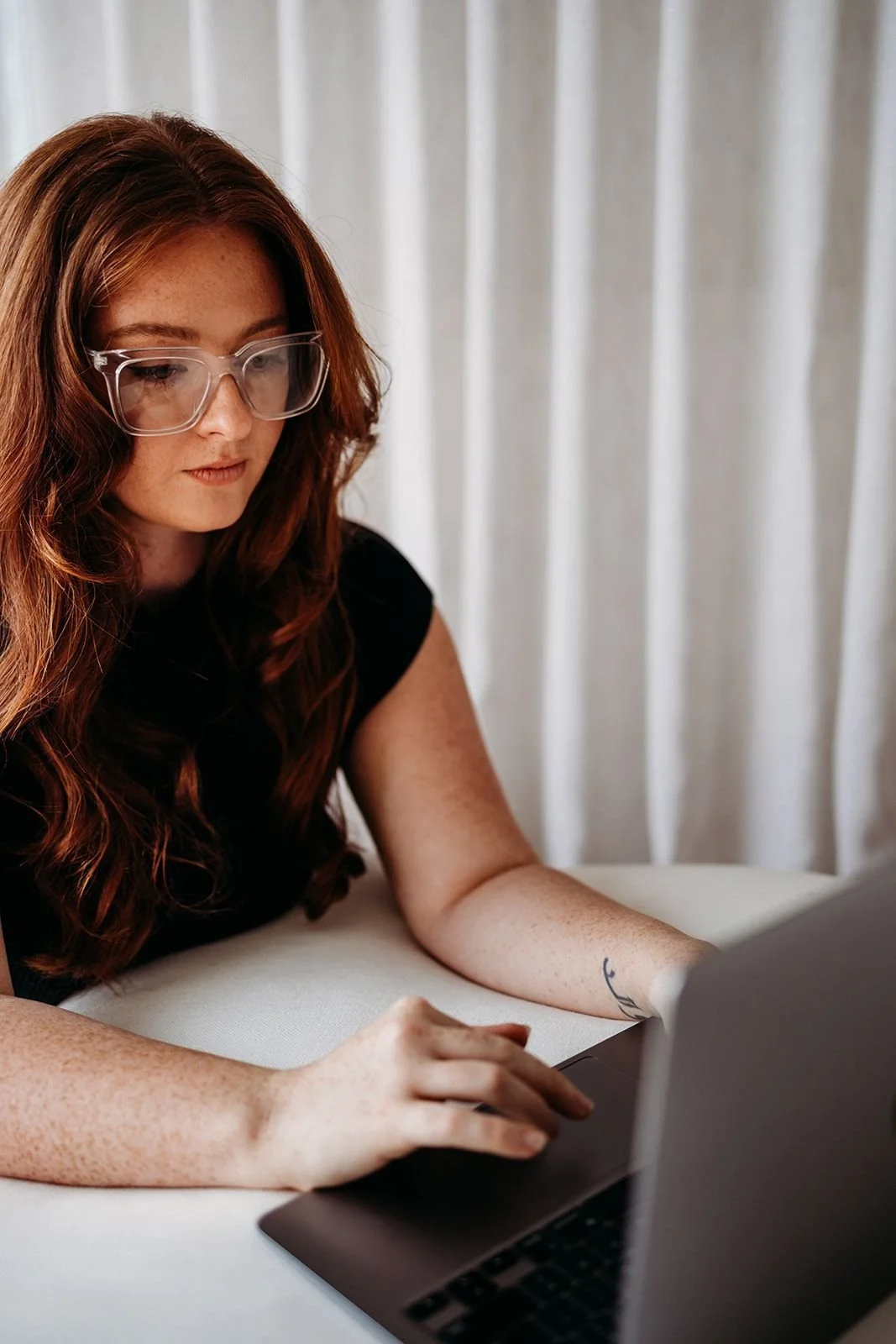 A young woman with long red hair and glasses working on a laptop at a table, with white curtains in the background.