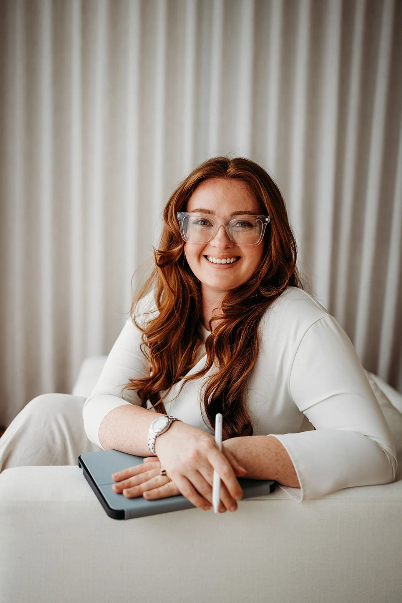 A woman with long red hair, wearing glasses and a white blouse, smiling while sitting at a table with a notebook and a pen in hand, against a background of beige curtains.