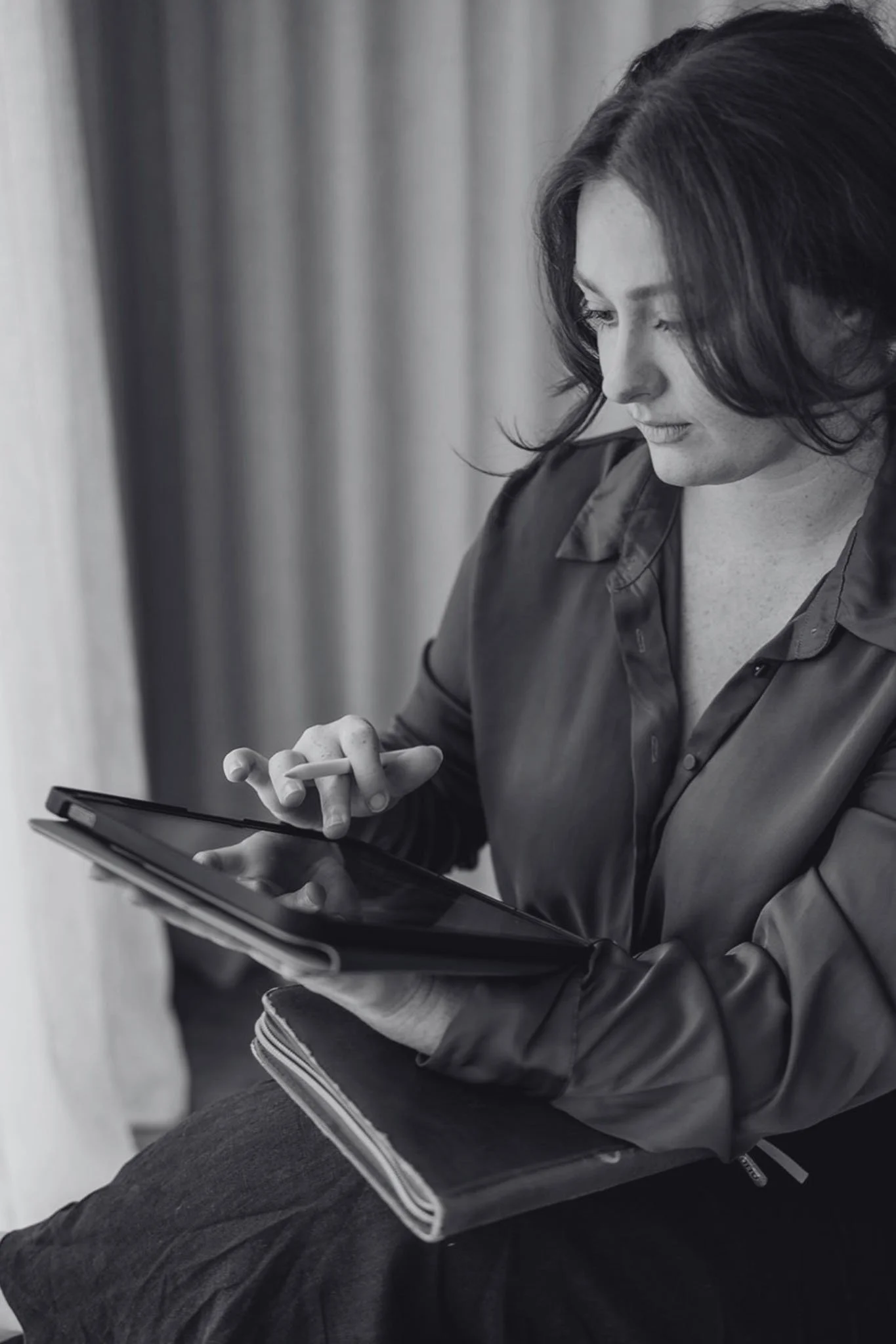 A woman with shoulder-length dark hair looking down at her phone while seated. She is wearing a dark jacket and sitting against a blurred background.