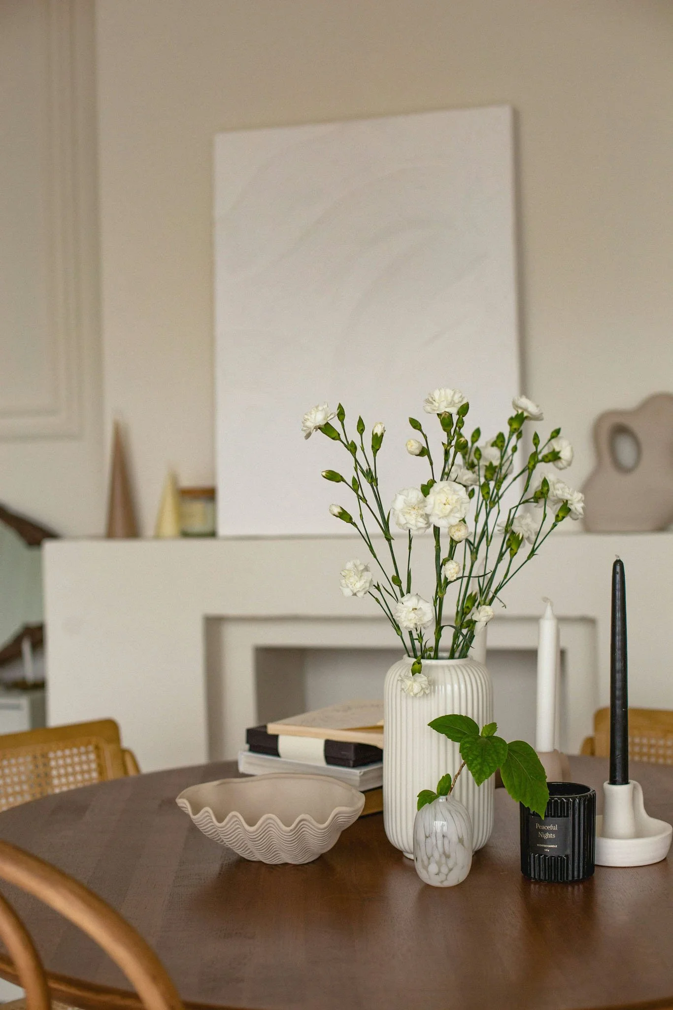 A dining table decorated with a white vase holding white flowers, a smaller white marble vase with green leaves, and various candles. There are also some books and a decorative white bowl on the table. In the background, a white wall with a blank canvas or whiteboard and some blurred decor items.