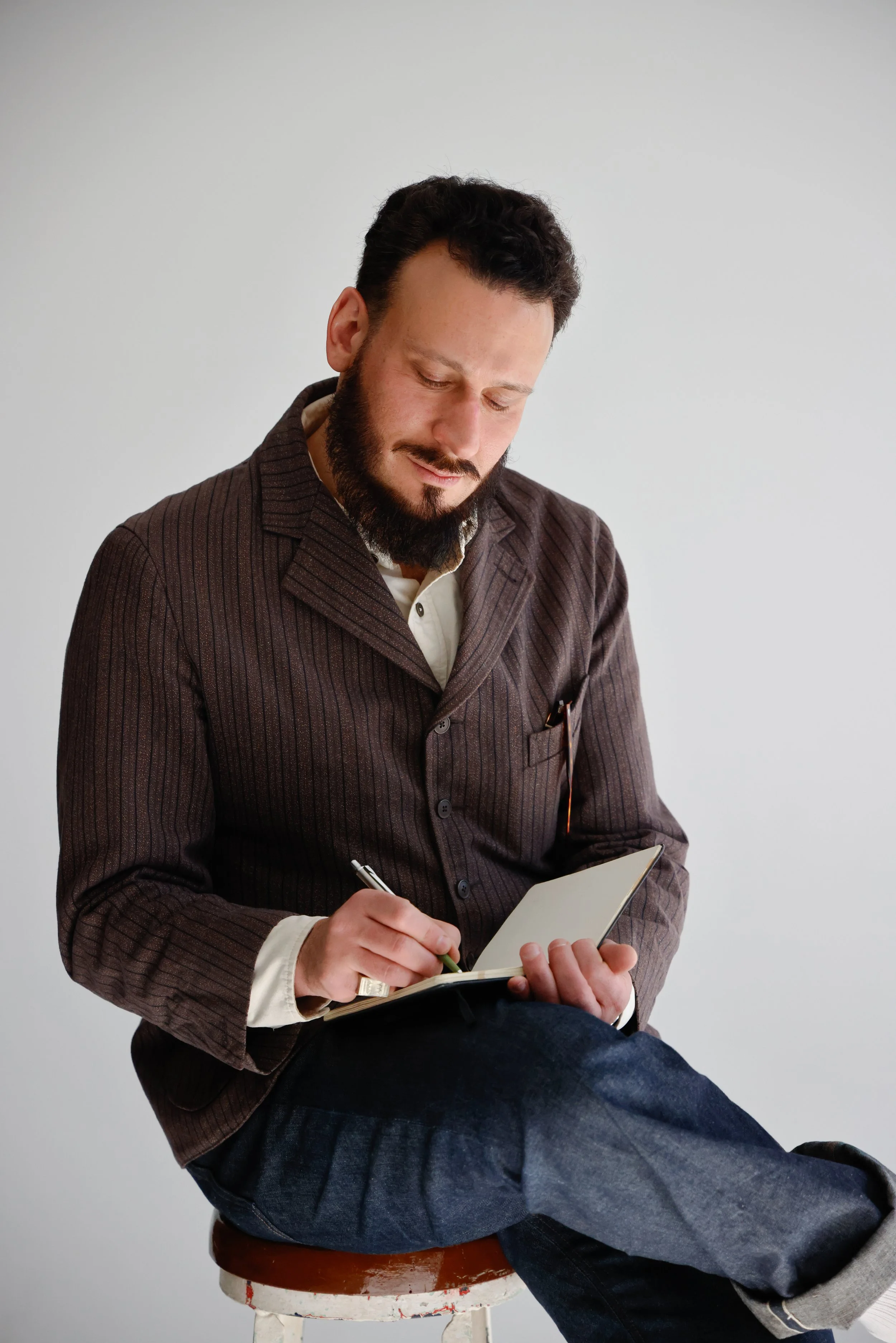 Man with dark hair and beard sitting on a stool, wearing a brown pinstripe blazer and blue jeans, writing in a notebook against a plain white wall.