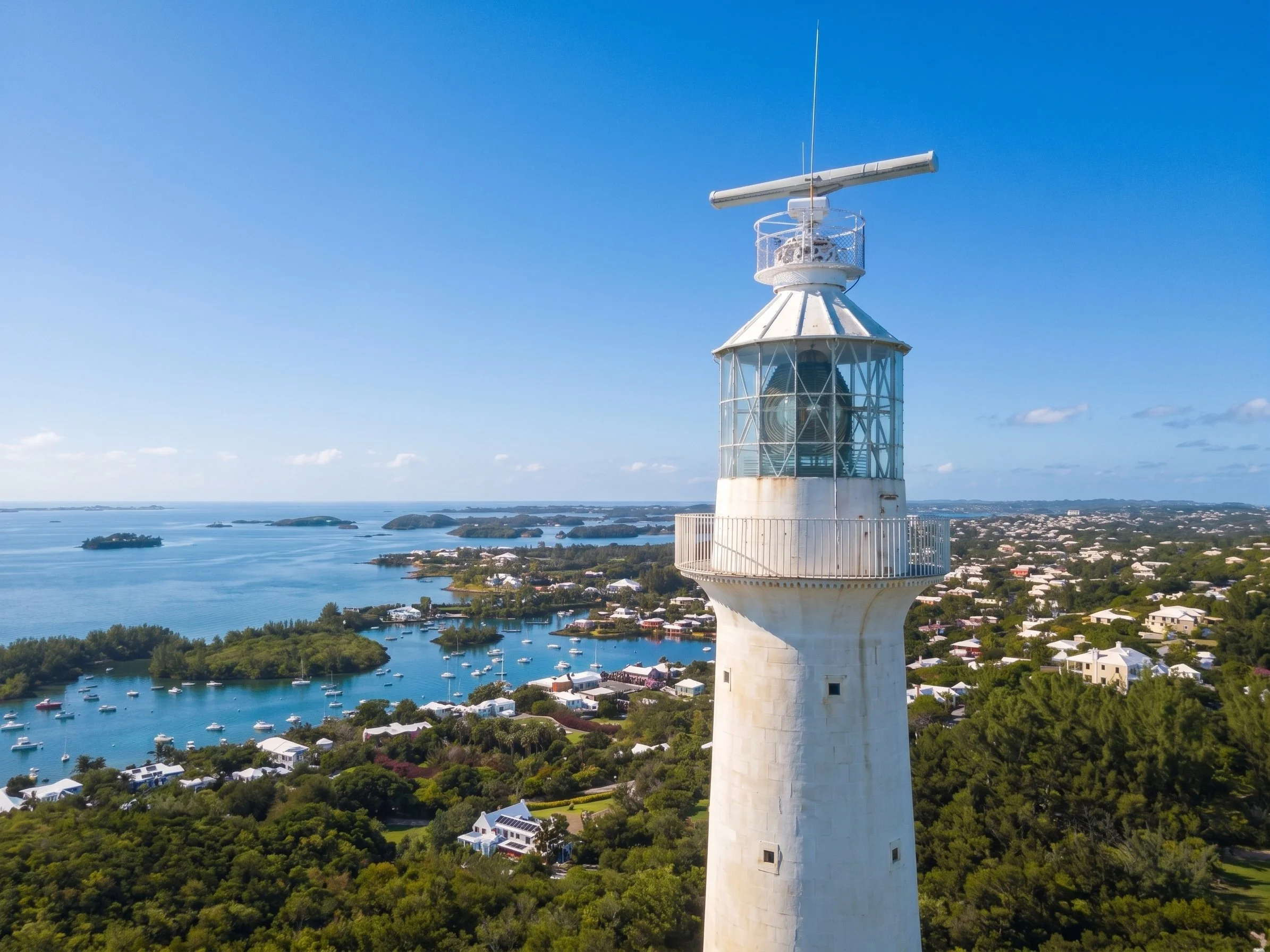 A white lighthouse with a glass-enclosed top and an antenna on top, overlooking a body of water with boats and islands, under a blue sky.