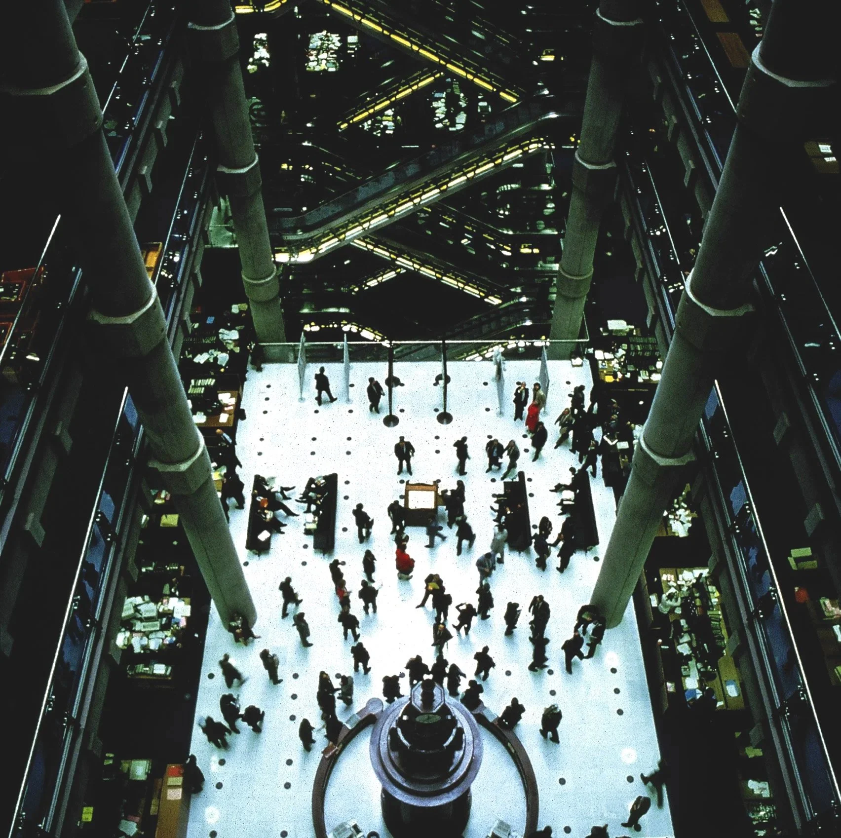 Interior view of a multi-level building with escalators, glass railings, people walking and shopping, and bright lighting.