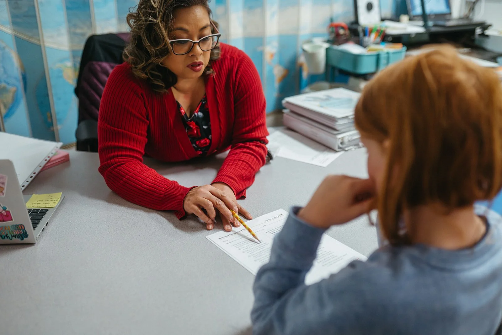 A woman in glasses and a red cardigan conducts a meeting with a young girl in a classroom or office setting. The woman points to a document on the table with a yellow pencil. There are stacks of papers, a laptop, and various office supplies on the desk.