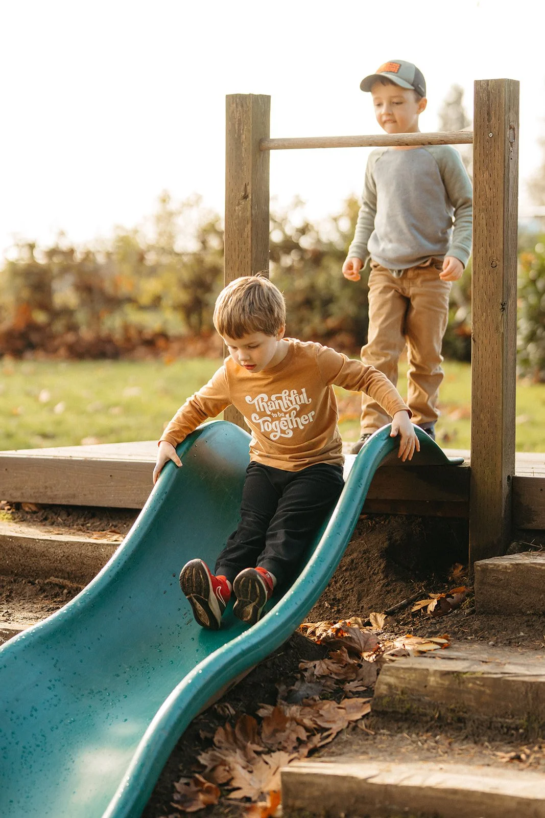 Two young boys play on a blue slide outdoors in autumn, one sliding down while the other stands at the top, surrounded by fallen leaves.