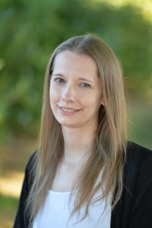 A young woman with long blonde hair, blue eyes, and fair skin outdoors with greenery in the background, smiling slightly at the camera.