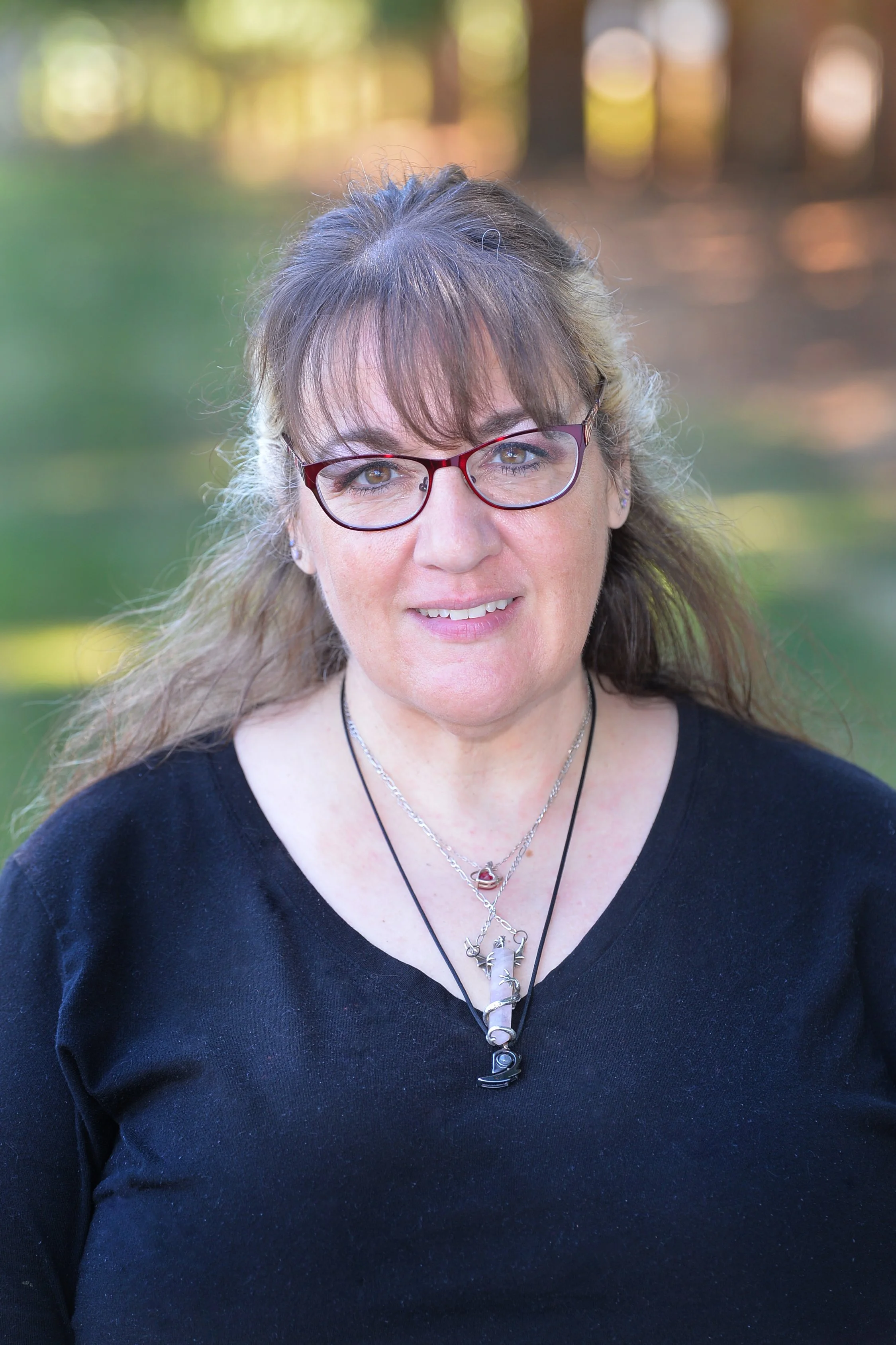 Close-up of a woman with brown hair, glasses, and wearing layered necklaces, outdoors with blurred trees in background.