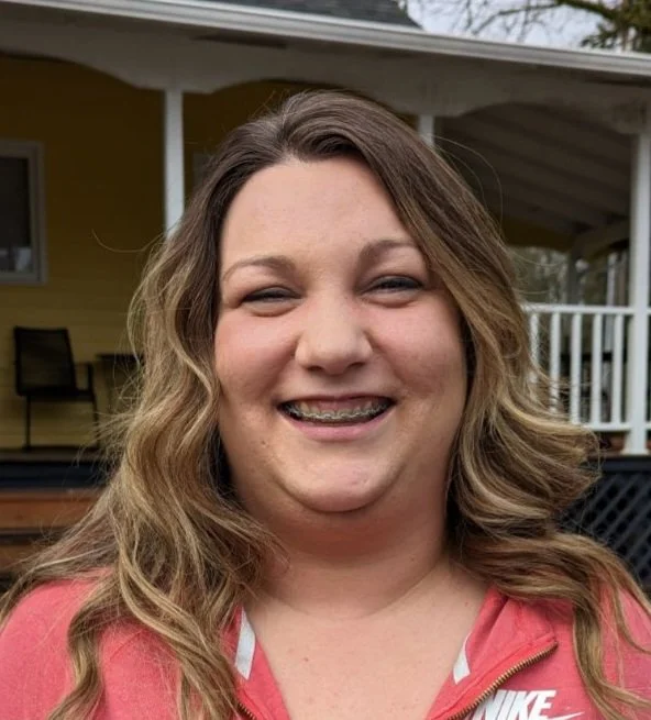 A woman with long wavy hair smiling outside in front of a yellow house with a porch and white railing, wearing a pink Nike zip-up jacket.