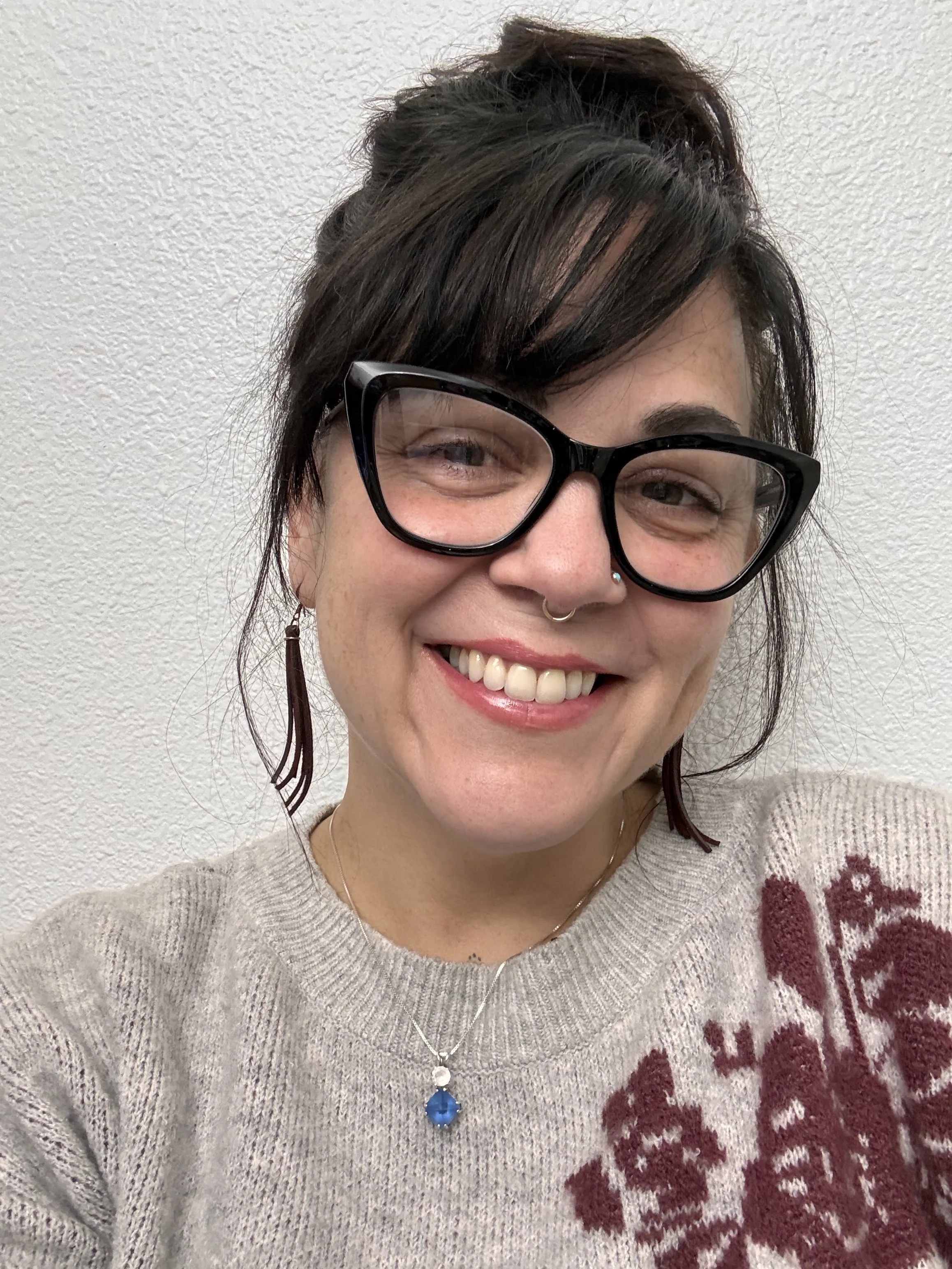 A woman with short dark hair, wearing large black glasses, earrings, a nose piercing, a necklace with a large blue pendant, and a beige sweater with red lettering, smiling in front of a textured white wall.