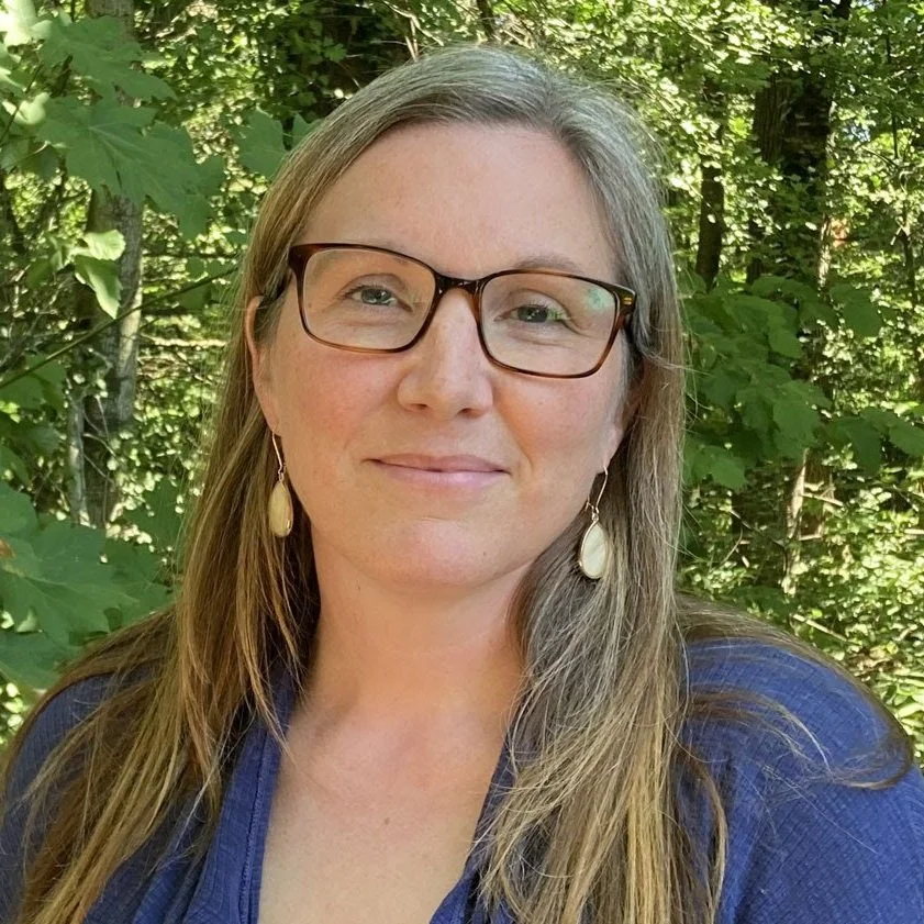 A woman with long hair and glasses standing outdoors in front of green leafy trees.