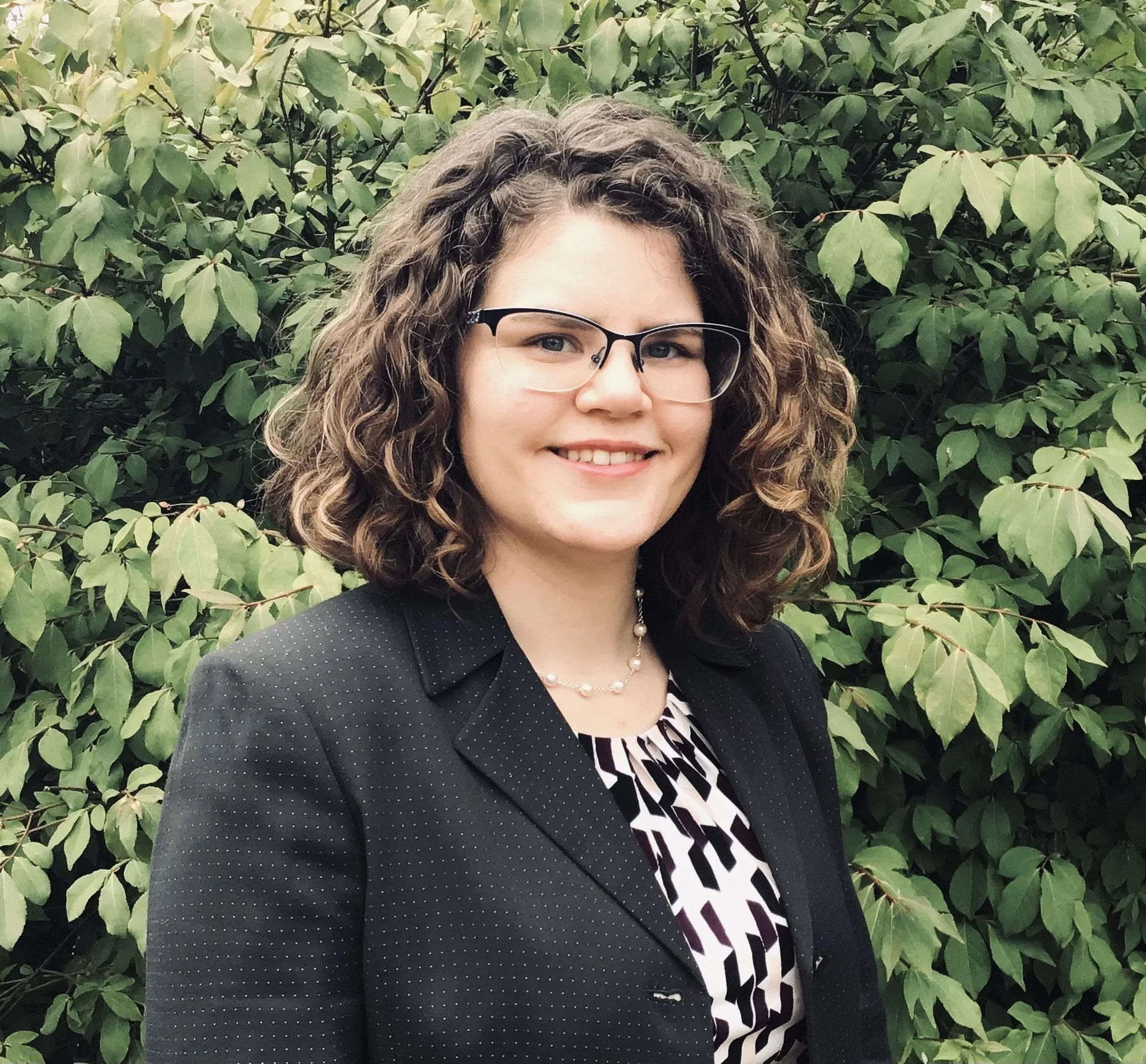 A smiling woman with curly brown hair and glasses standing in front of green leafy bushes, wearing a black blazer with small white dots, a patterned blouse, and a pearl necklace.