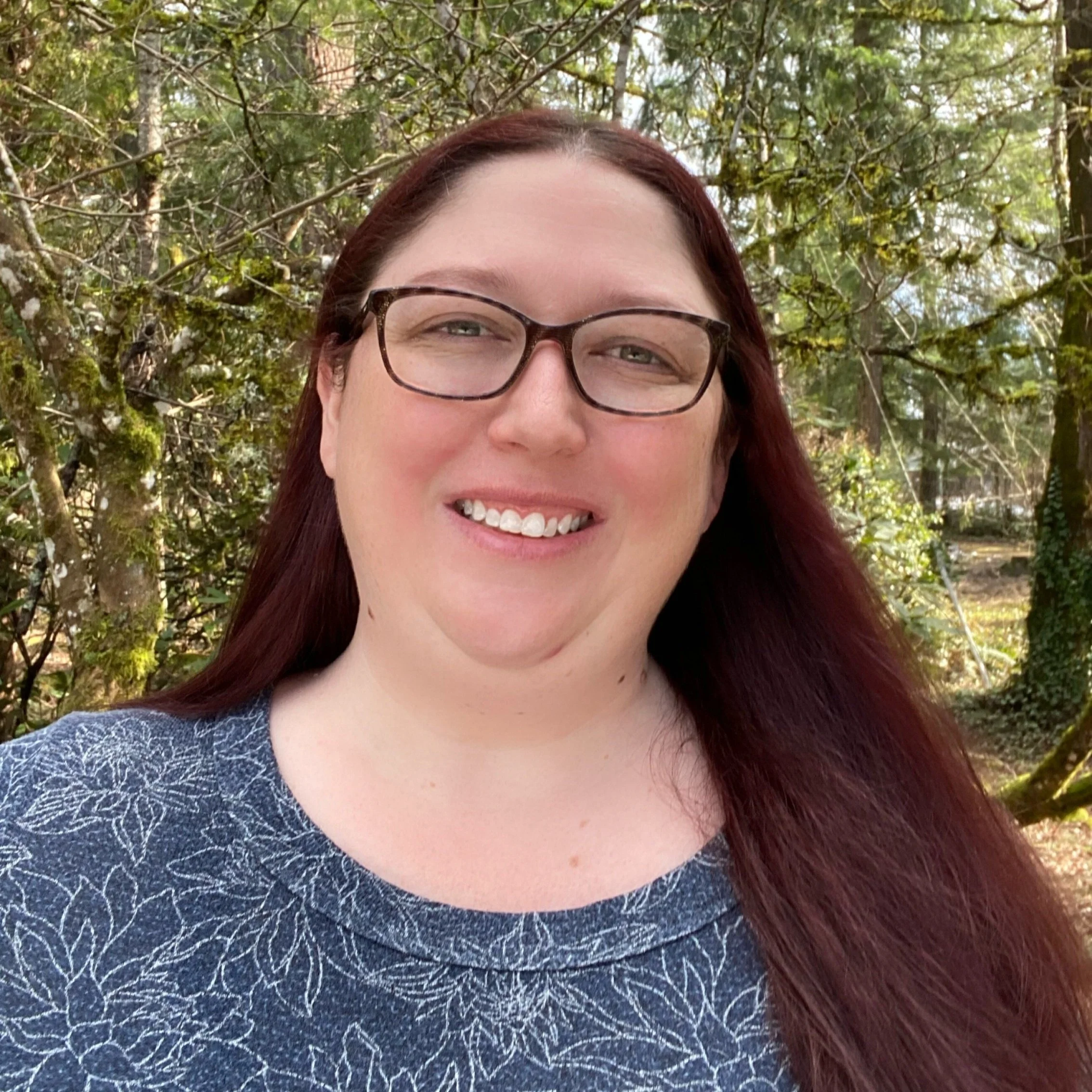 A woman with glasses and red hair smiling outdoors, with a background of trees and foliage.