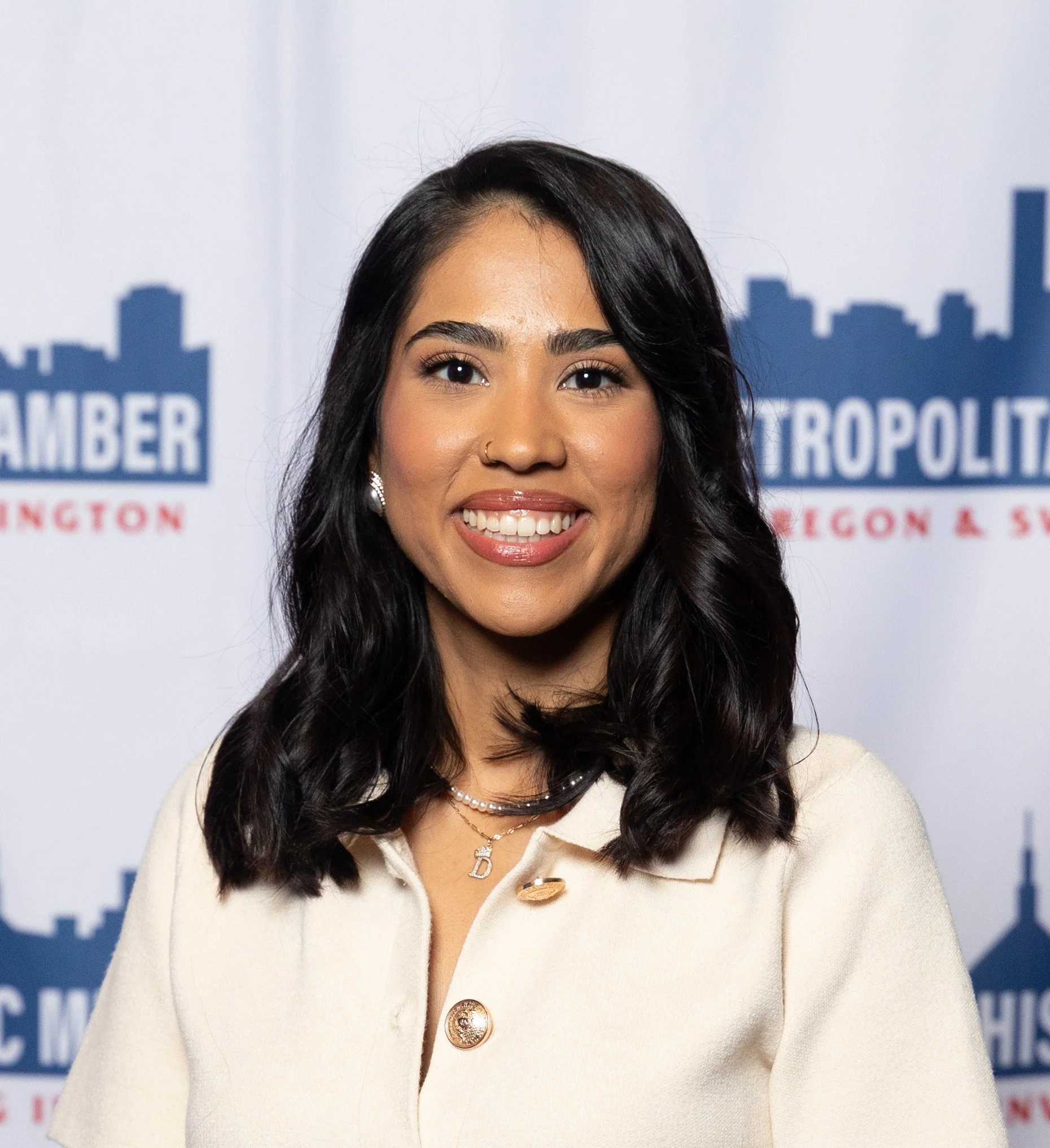 Close-up of a young woman with black hair and brown eyes smiling, wearing a cream-colored blazer with large gold buttons, layered pearl necklaces, and a nose ring, standing in front of a backdrop with a cityscape and the words 'Chamber of Commerce & Industry'.