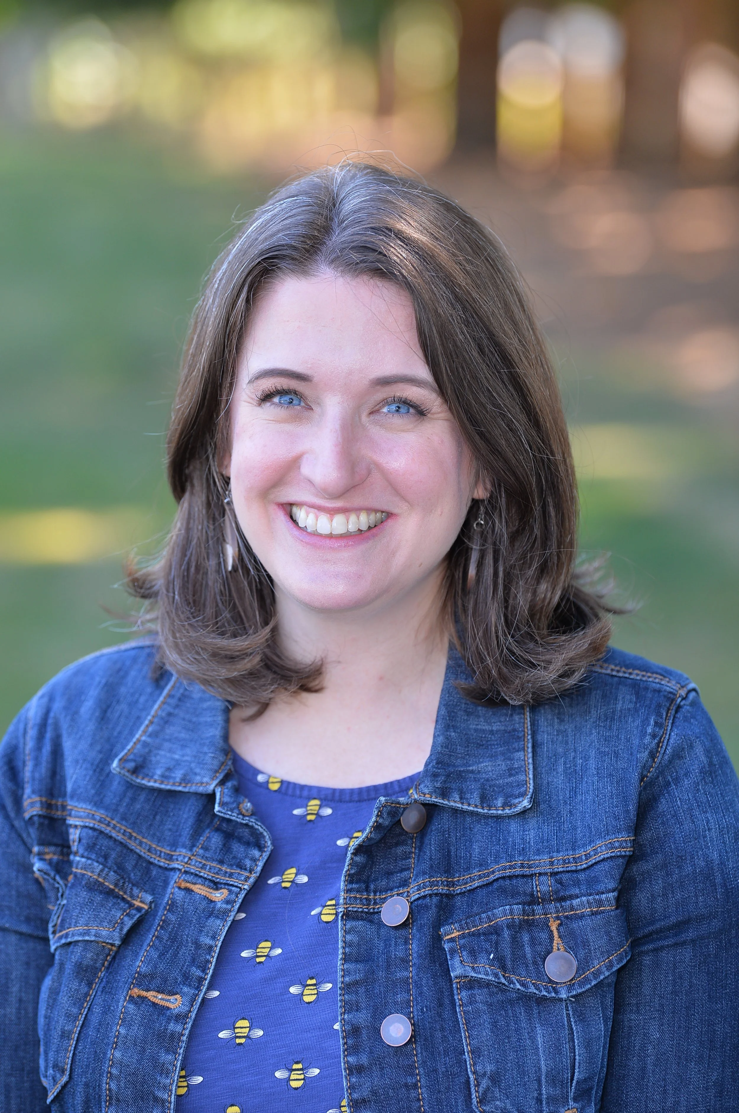 Smiling woman with shoulder-length brown hair, wearing a denim jacket over a blue shirt with yellow bee pattern, standing outdoors with blurred background of trees and sunlight.