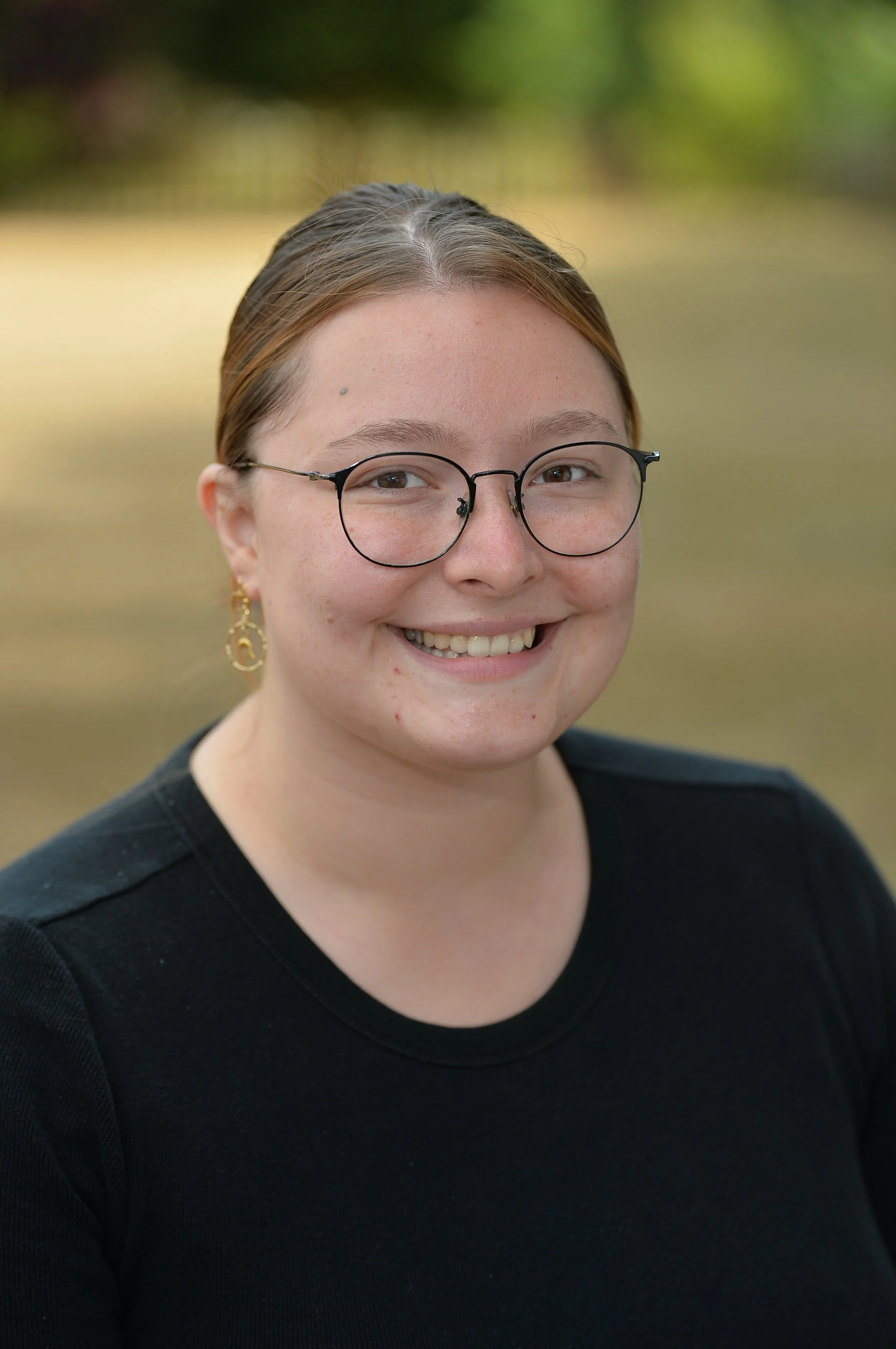 Close-up of a young woman with glasses, earrings, smiling outdoors with greenery in the background.
