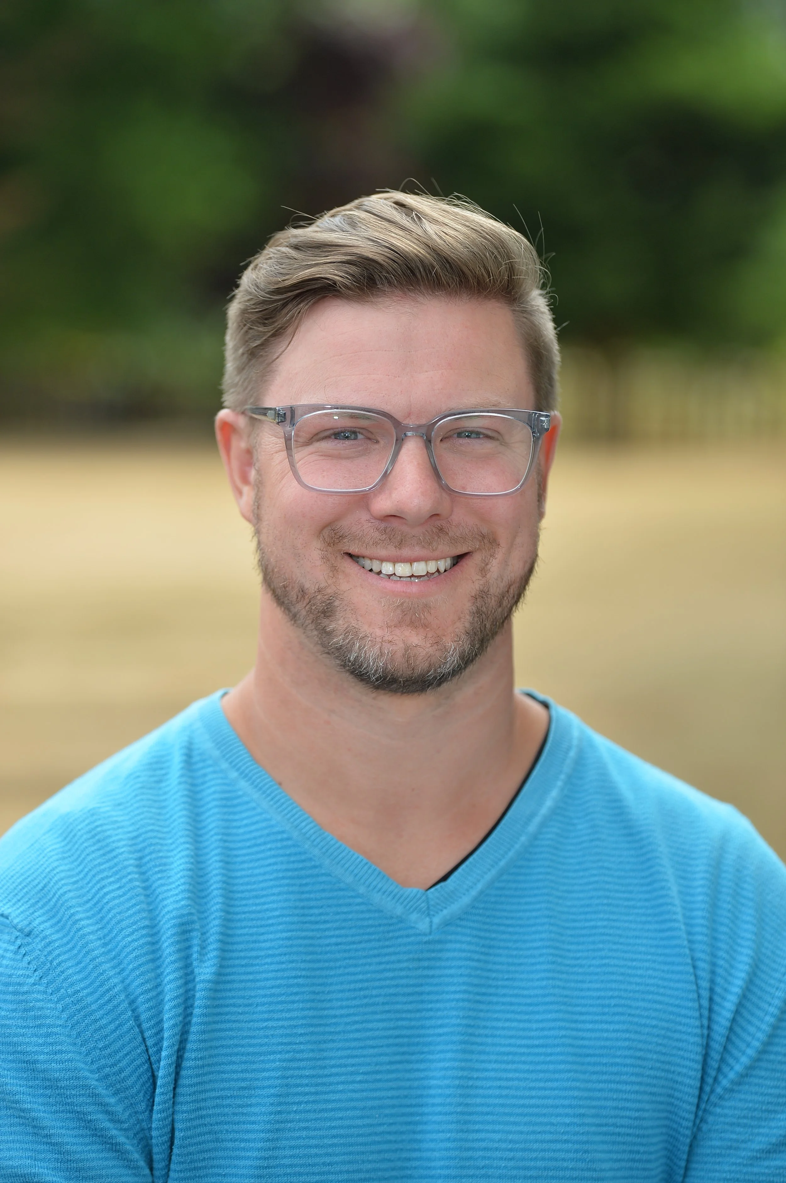 Close-up of a smiling man wearing clear glasses and a light blue V-neck shirt, outdoors with a blurred background of green trees and yellow grass.