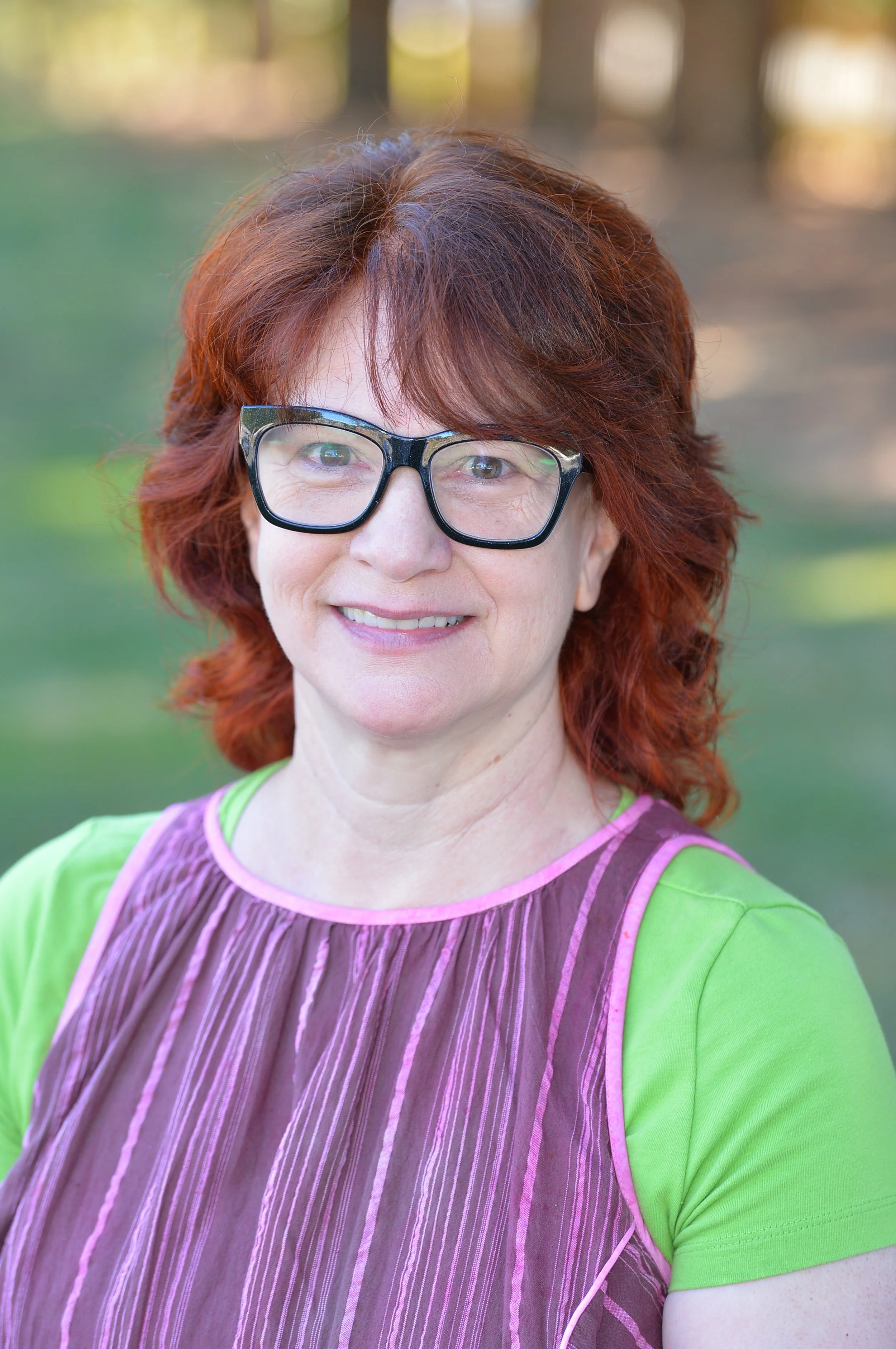 A woman with red hair, glasses, and a bright smile outdoors during daytime. She is wearing a green shirt and a brown and pink striped vest.