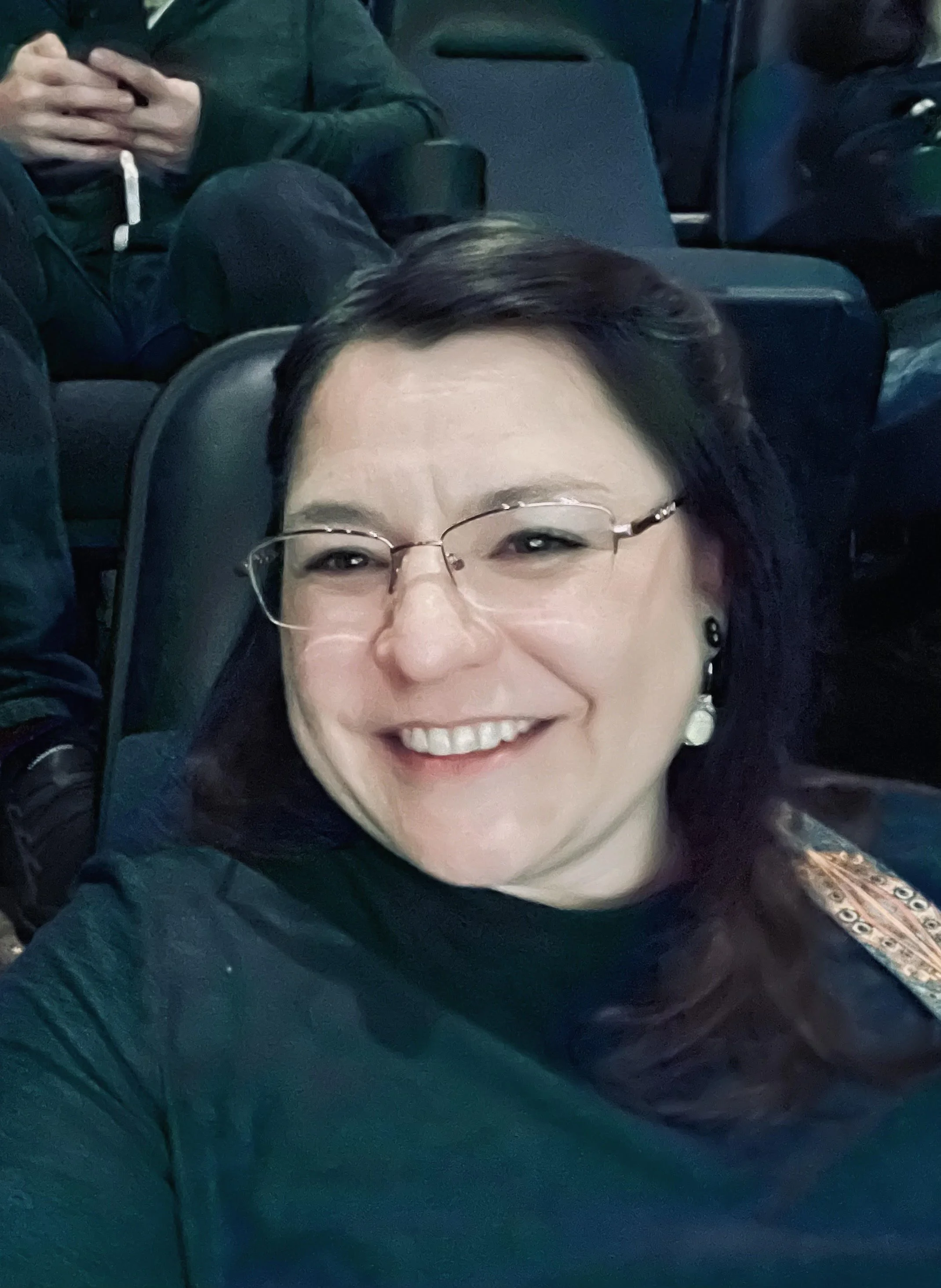 Close-up of a smiling woman with glasses, dark hair, and earrings, wearing a dark jacket, inside an airplane.