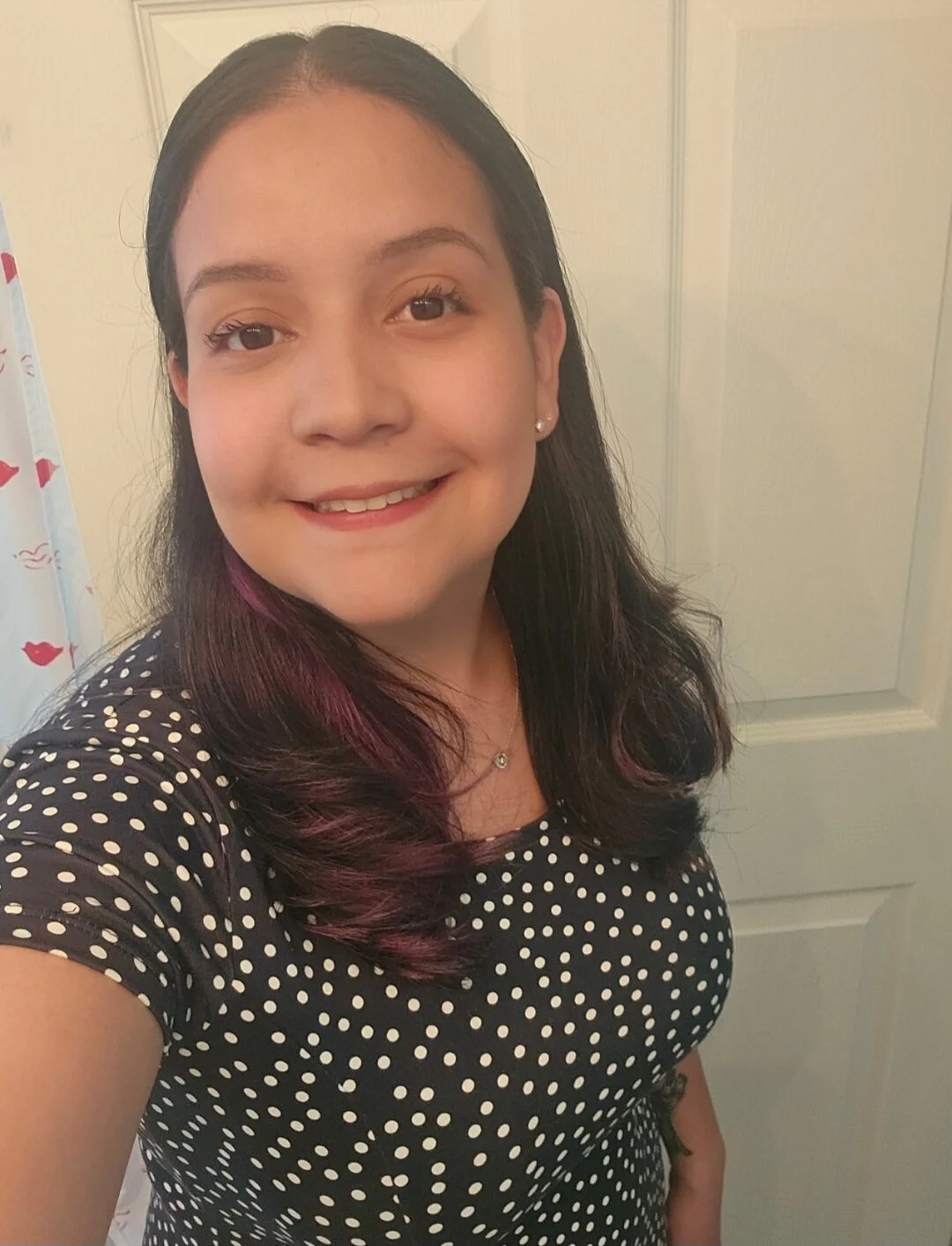 A young woman with long dark hair streaked with purple, smiling and taking a selfie in front of a plain door.