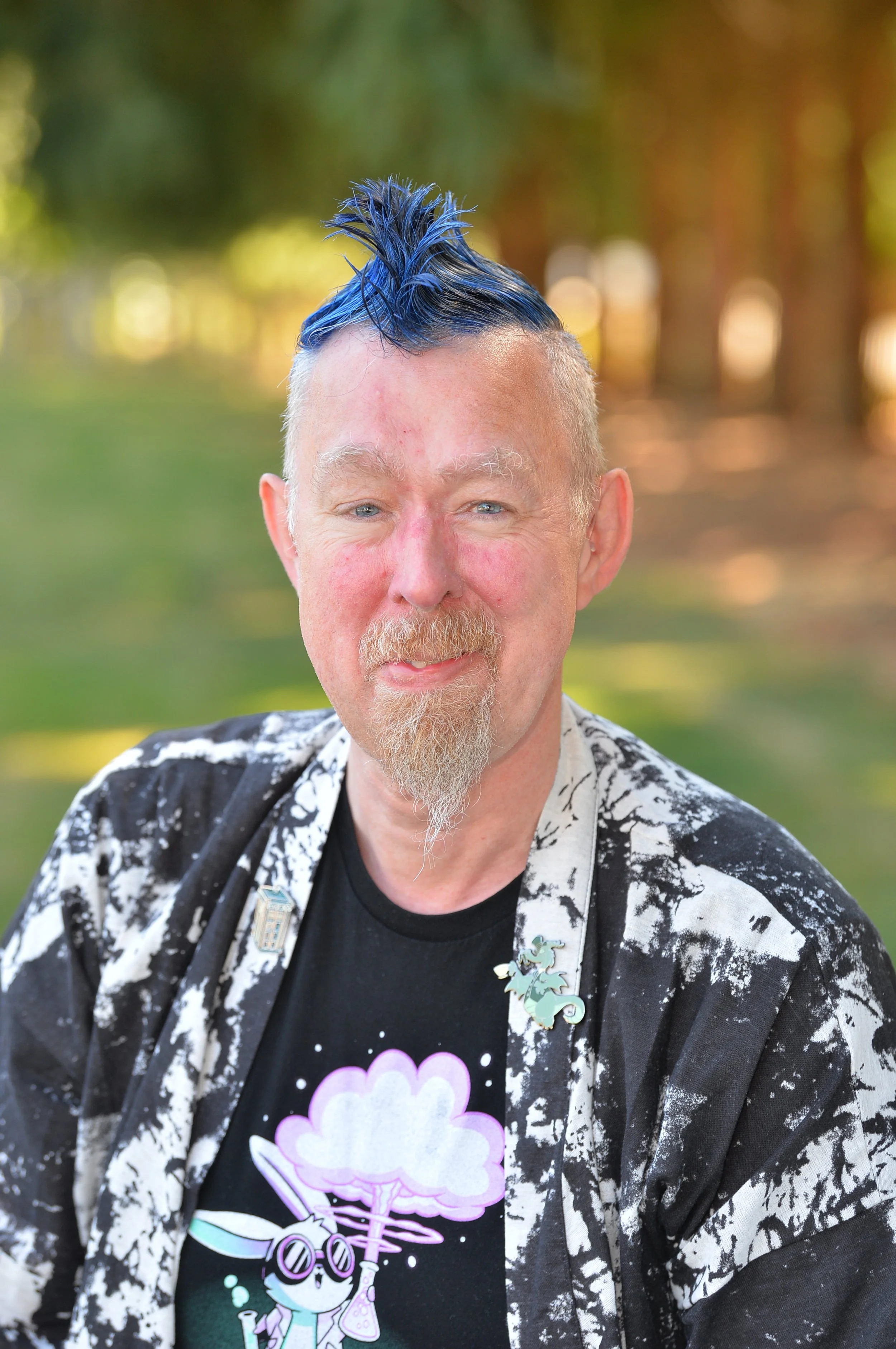 A man with blue hair styled in a spiky mohawk, fair skin, and blue eyes outdoors with blurred trees in the background. He is wearing a black t-shirt with a cartoon kitten in a lab coat and glasses, and a black and white patterned jacket with various pins and patches.