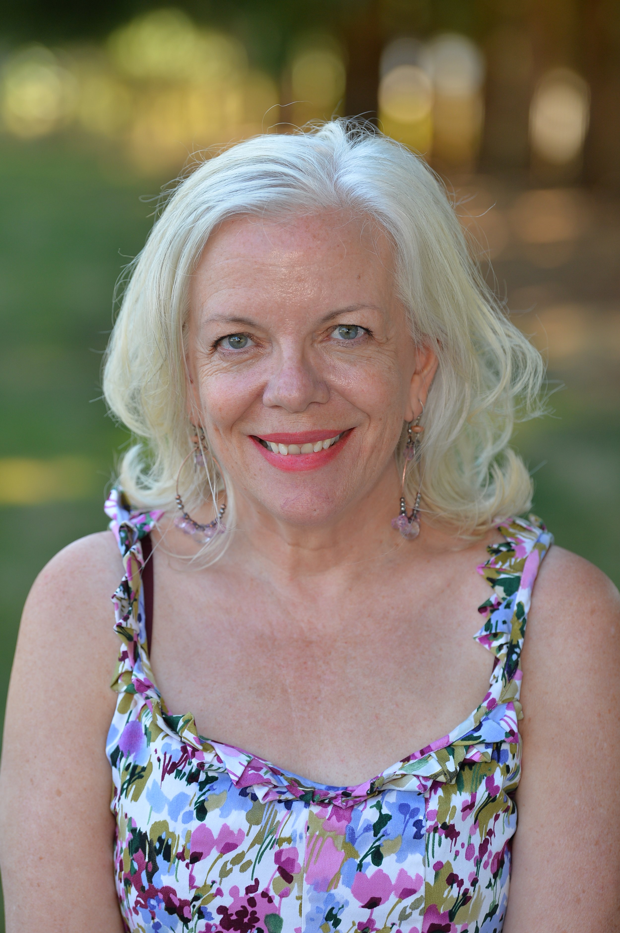 A smiling elderly woman with shoulder-length white hair and blue eyes, wearing a floral sleeveless dress with purple, pink, and green colors, outdoors with a blurred background of trees and sunlight.