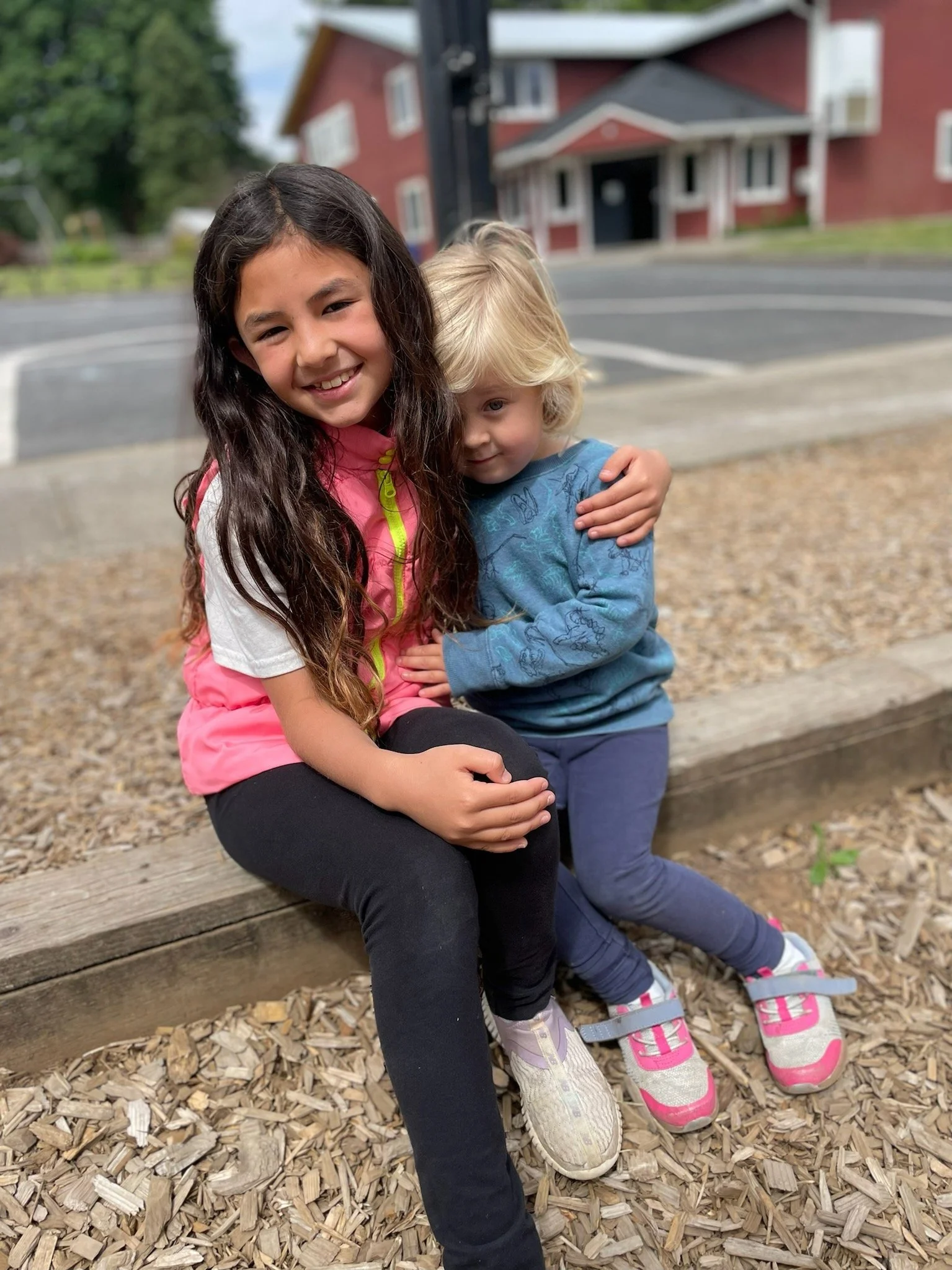 Two children, a girl with long dark hair in a pink vest and a boy with blonde hair in a blue shirt, sitting on a wooden ledge outdoors, hugging each other with a house and trees in the background.