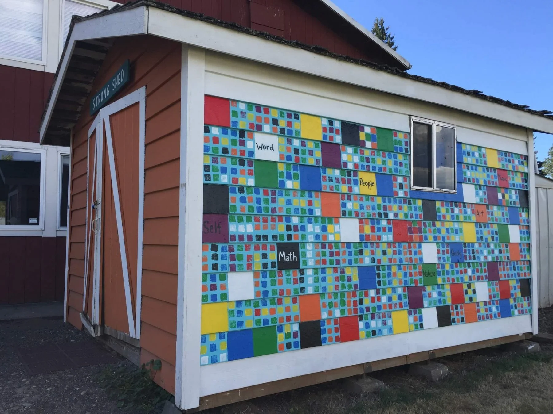 A small building with a colorful mosaic on the wall, labeled with words like 'Word,' 'People,' 'Art,' and 'Math.' The building has brown and white siding, a window, and a sign that reads 'Strong shed.'