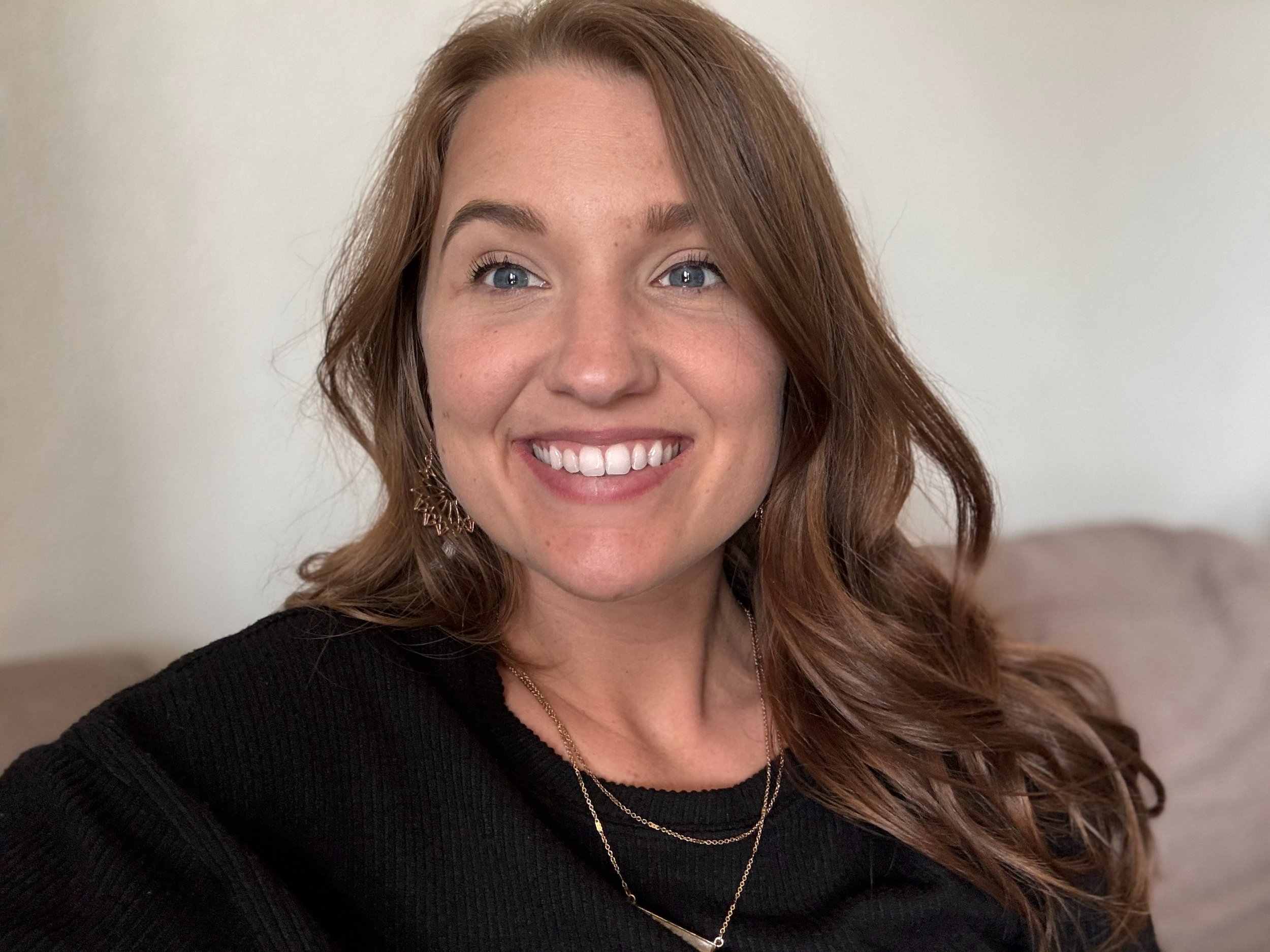 Close-up of a smiling woman with long, wavy, reddish-brown hair, blue eyes, wearing a black top and gold jewelry, standing against a plain white wall.