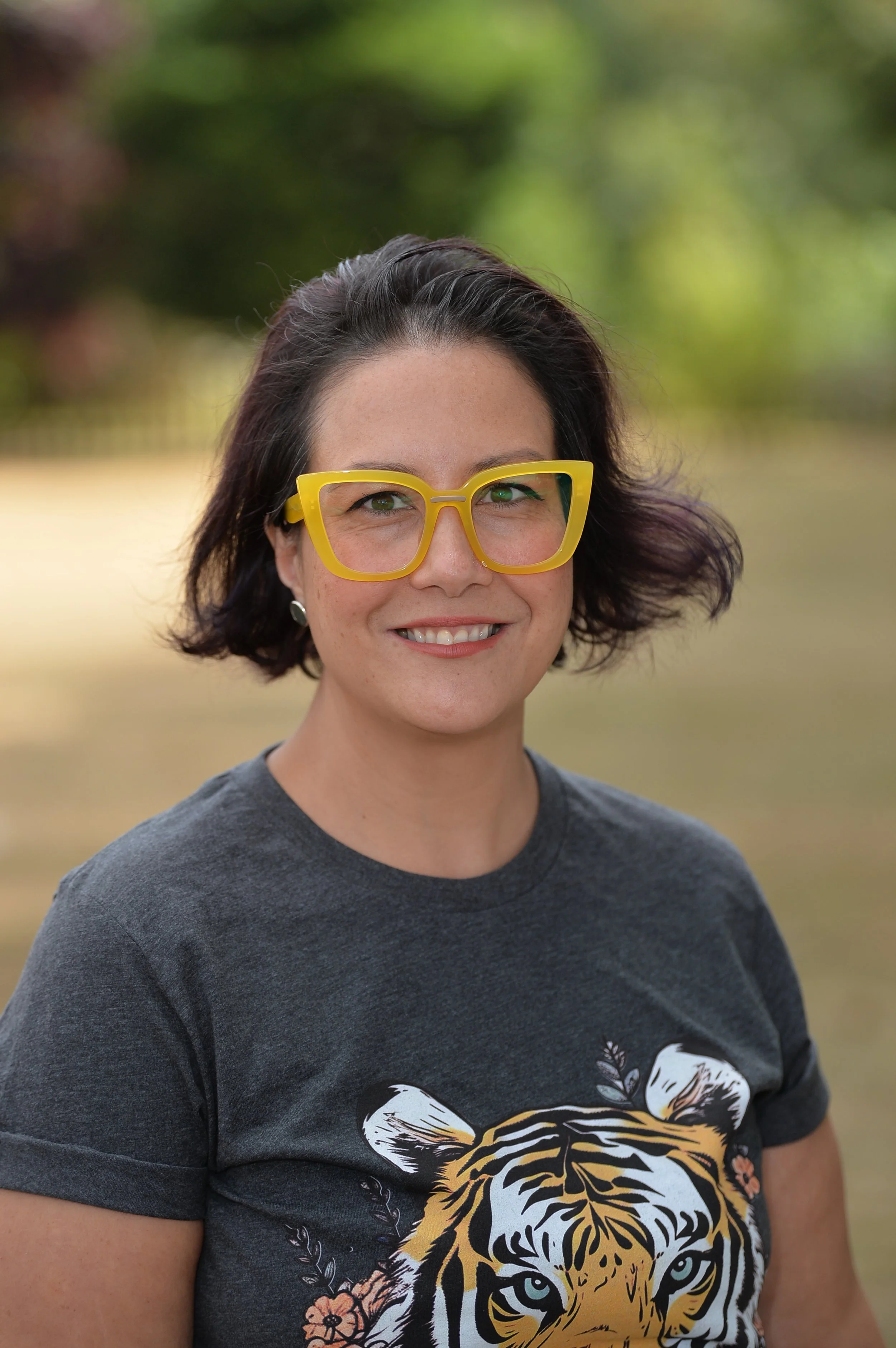 A woman with short dark hair wearing large yellow glasses and a dark T-shirt featuring a tiger's face, outdoor background with blurred green trees.