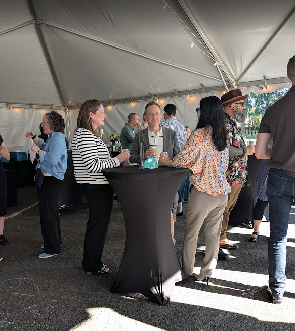 People gathered under a large tent at a social event, standing around high tables, chatting and drinking wine.
