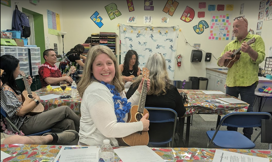 People in a classroom playing ukuleles while a man plays guitar at the front. One woman is smiling at the camera. The room is decorated with colorful signs and posters.