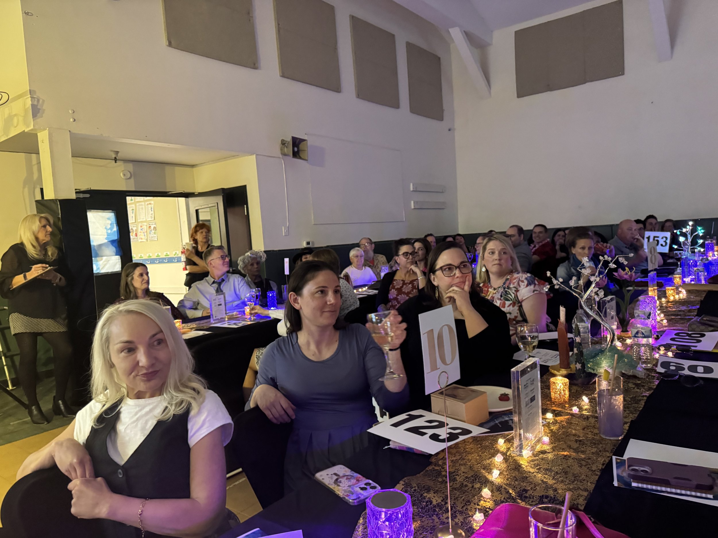 A crowded banquet hall with people sitting at tables adorned with decorative centerpieces and candles, all facing toward the front where there is a white wall and a speaker setup, possibly for a presentation or speech.