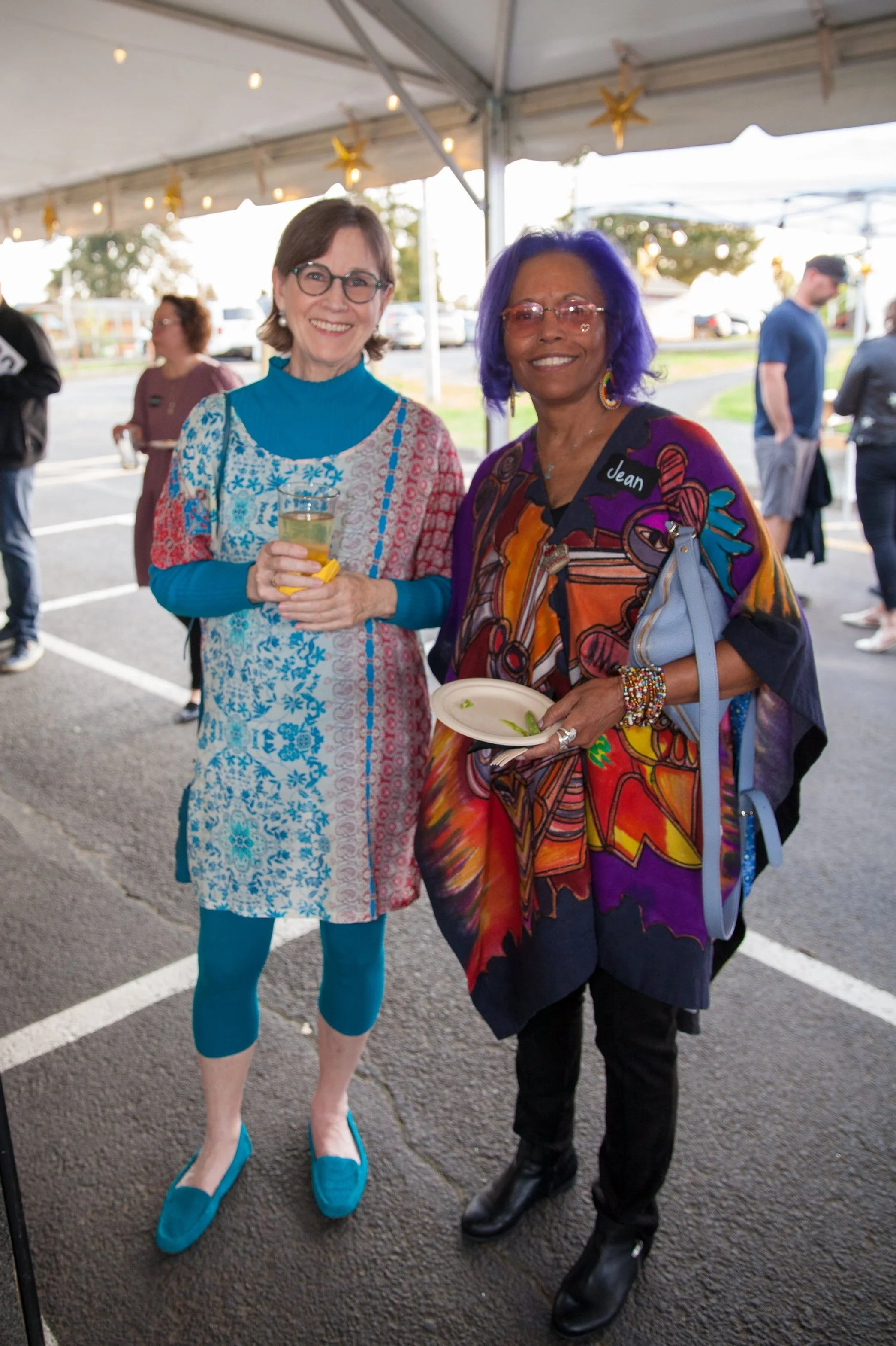 Two women standing under a tent at an outdoor event, smiling and posing for the photo. One woman is wearing a colorful dress with blue tights and matching shoes; the other is wearing a vibrant, patterned jacket with a black top, black pants, and blac