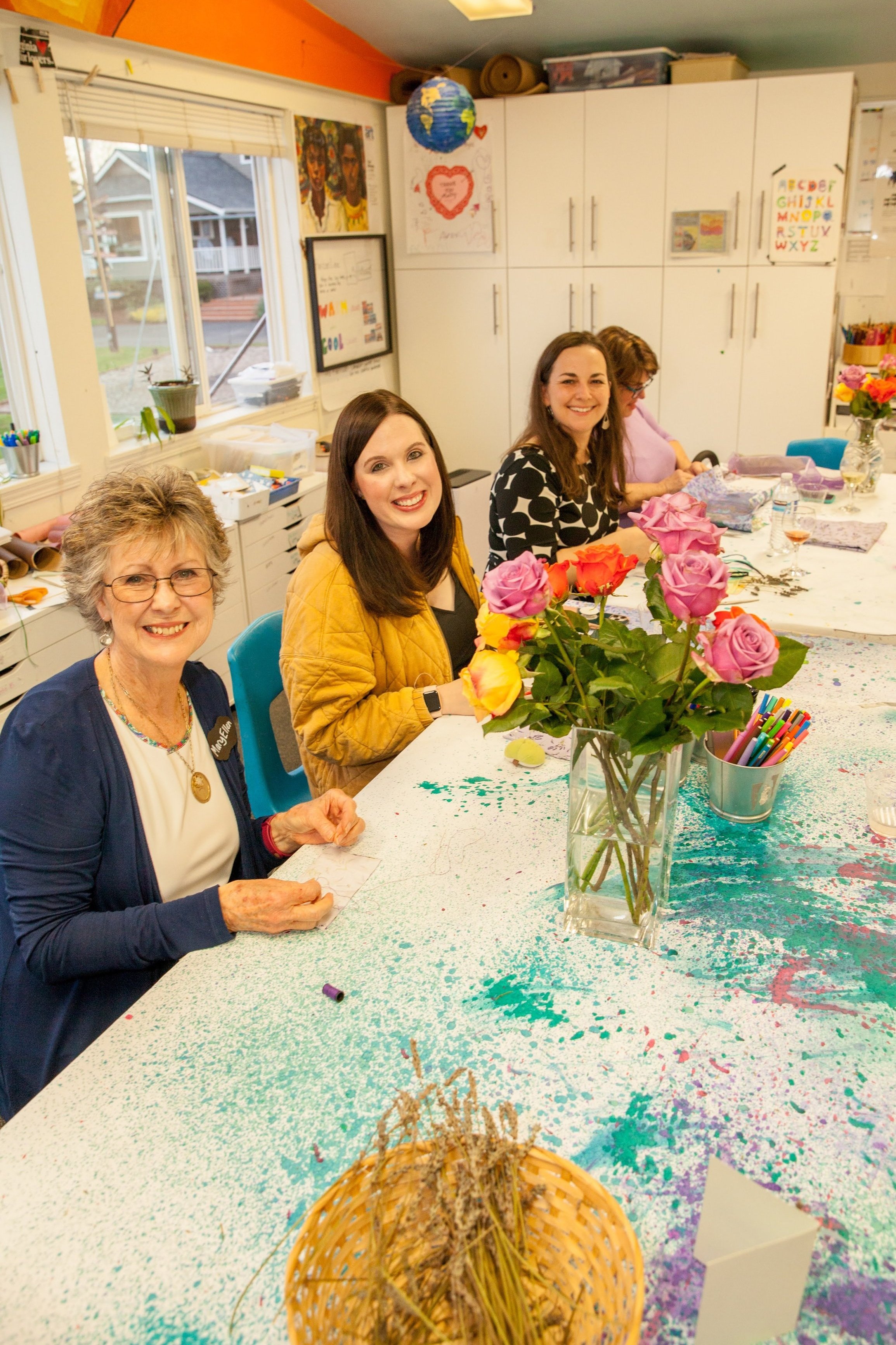 Four women sit around a table inside a colorful classroom decorated with artwork and alphabet charts. The table is decorated with a large vase of pink and orange roses. The tablecloth has multicolored paint splatters. One woman in a blue cardigan and
