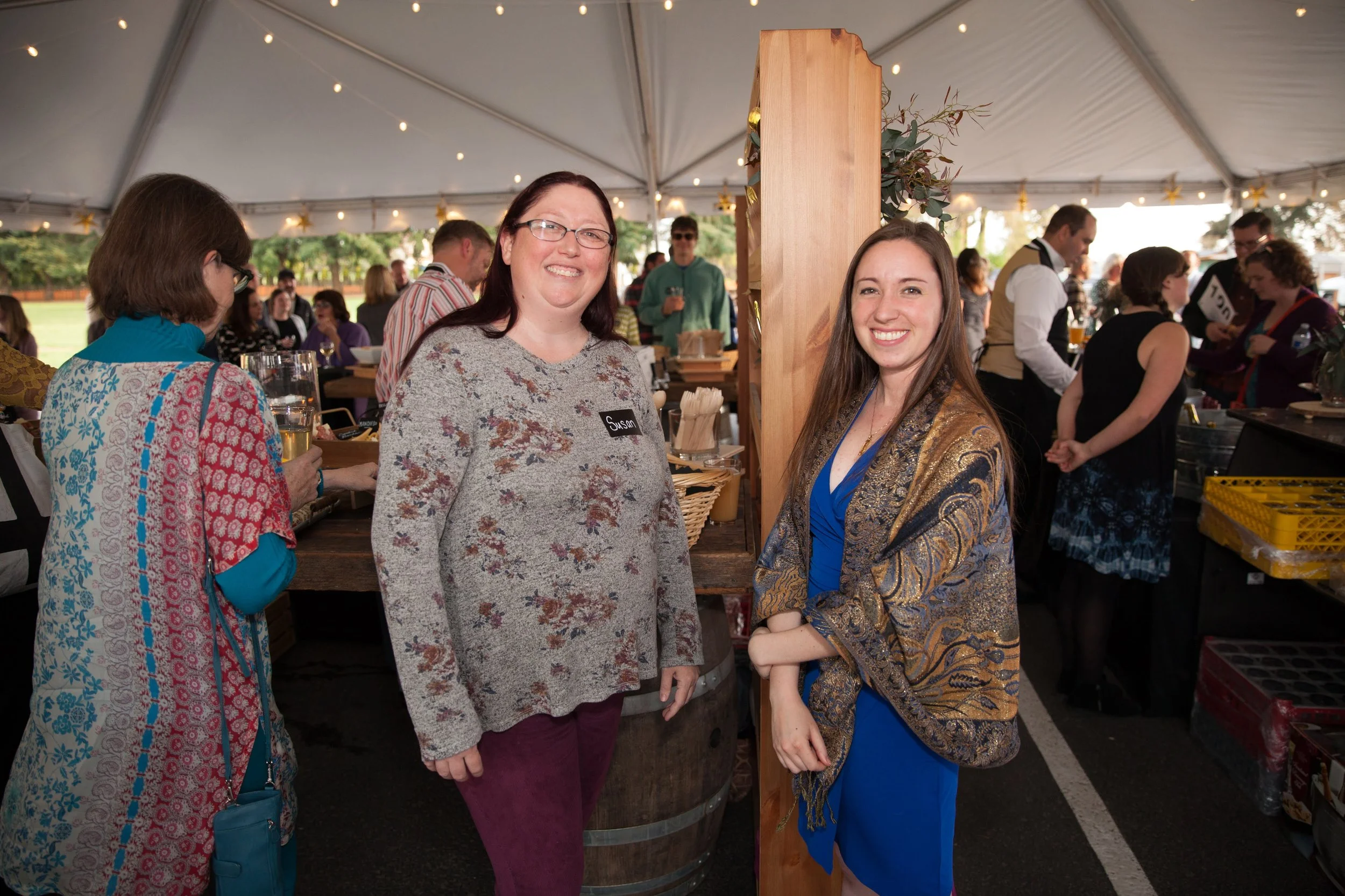 Two women standing together at a social gathering inside a large white tent, smiling at the camera. One woman is wearing glasses and a gray sweater, the other woman is in a blue dress with a patterned shawl. There are people in the background, some e