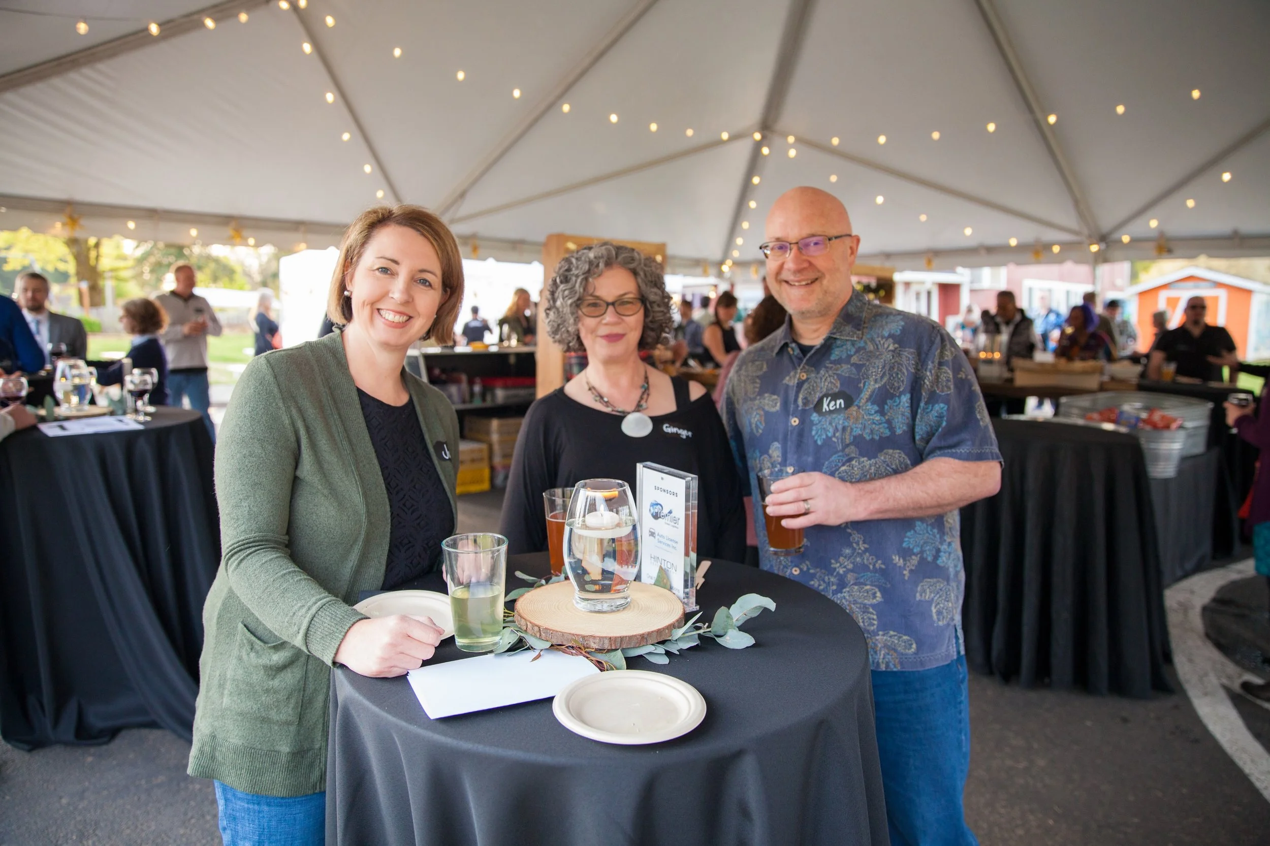Three people smiling at a table during an event under a large tent with string lights. The group includes a woman with short brown hair wearing a green blazer, a woman with curly gray hair and glasses wearing a black top, and a bald man with glasses 