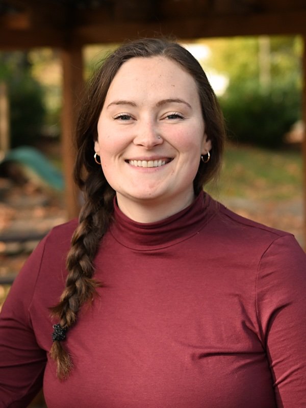 A smiling woman with long brown hair in a braid, wearing a maroon turtleneck, standing outdoors with blurred trees and structures in the background.